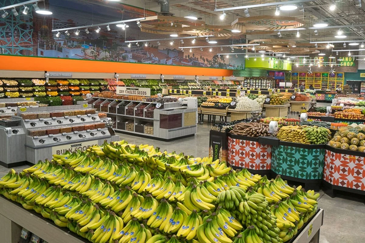 Interior of a grocery store with colorful produce displays, including bananas in the foreground, and various fruits and vegetables arranged in sections across the store.