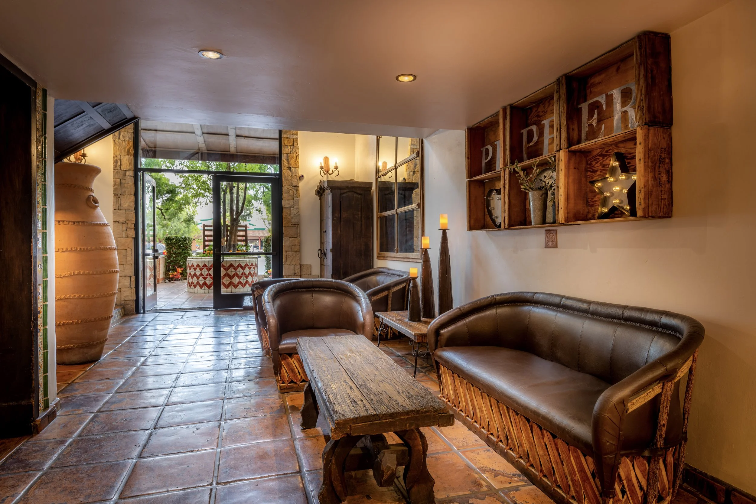 Rustic hotel lobby with leather sofas, a wooden coffee table, candleholder decorations, and glass door leading to an outdoor patio entrance with trees.
