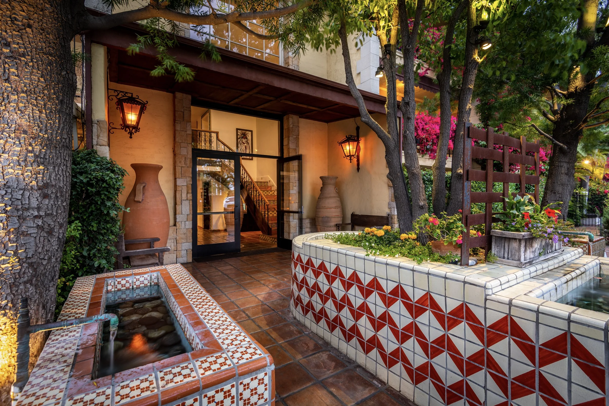 Outdoor patio with colorful tiles, potted plants, large clay vases, trees, and warm lighting.