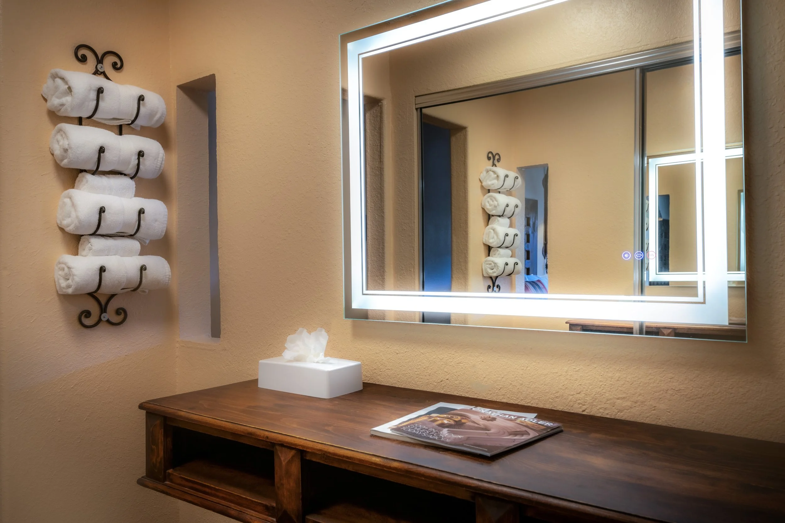 A bathroom vanity area with a illuminated rectangular mirror, a wooden tabletop holding a tissue box and a magazine, a wall-mounted towel rack with six rolled white towels, and beige painted walls.