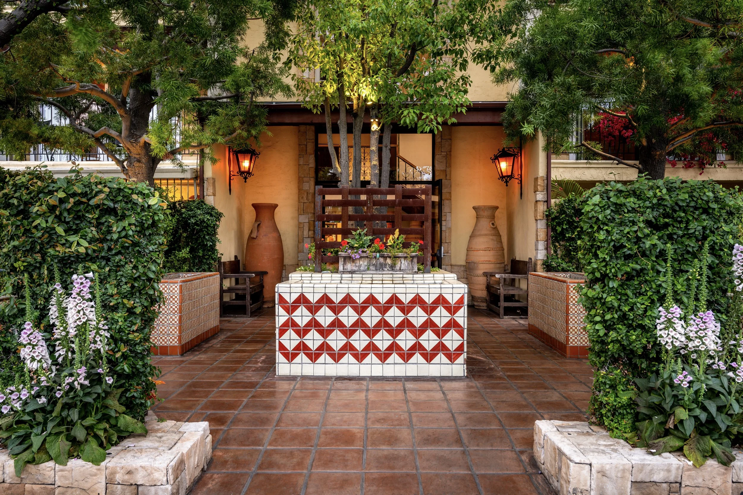 Outdoor patio with terracotta tiles, lush green bushes, flowering plants, and two large clay pots, featuring an entryway with warm lighting and decorative tiles.