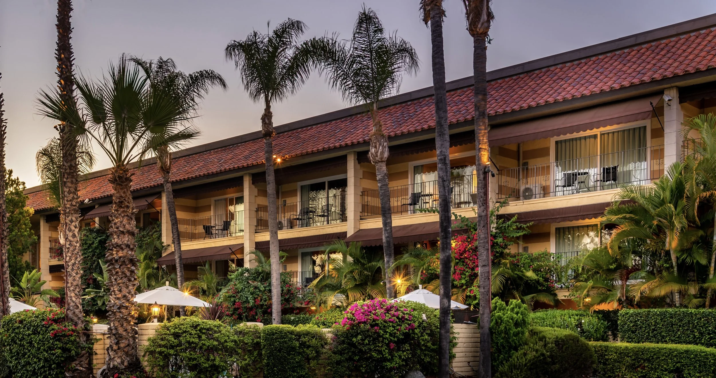 Landscape view of a multi-story hotel with balconies, pink flowering bushes, lush green plants, and tall palm trees at sunset.