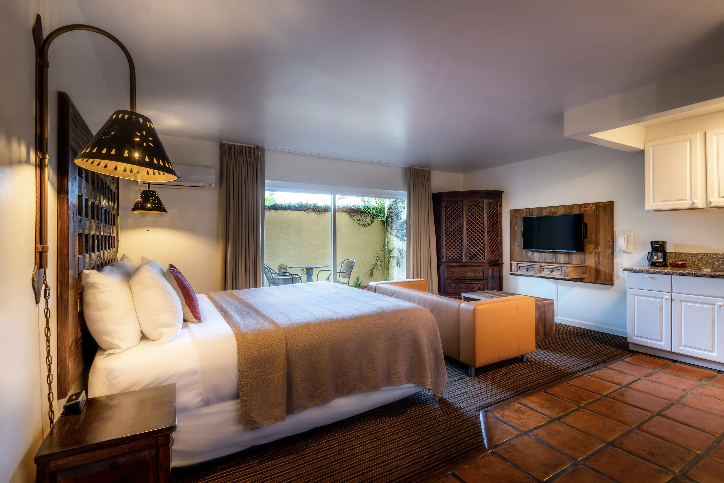 Hotel room with a large bed, bedside table, mounted TV, small kitchen area, and sliding glass door leading to a patio with outdoor seating.