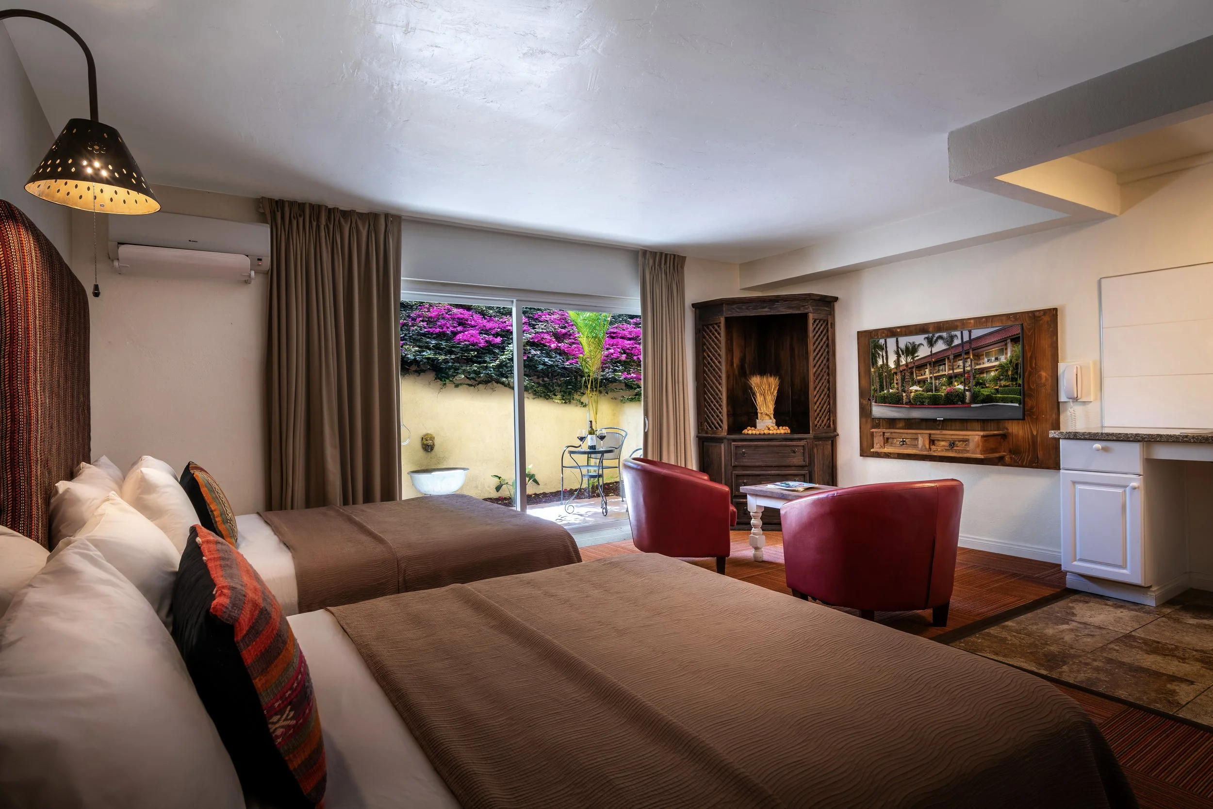 Hotel room with two beds, a sliding glass door leading to a garden, a wall-mounted TV, two red chairs, a small table, and a corner wooden cabinet.