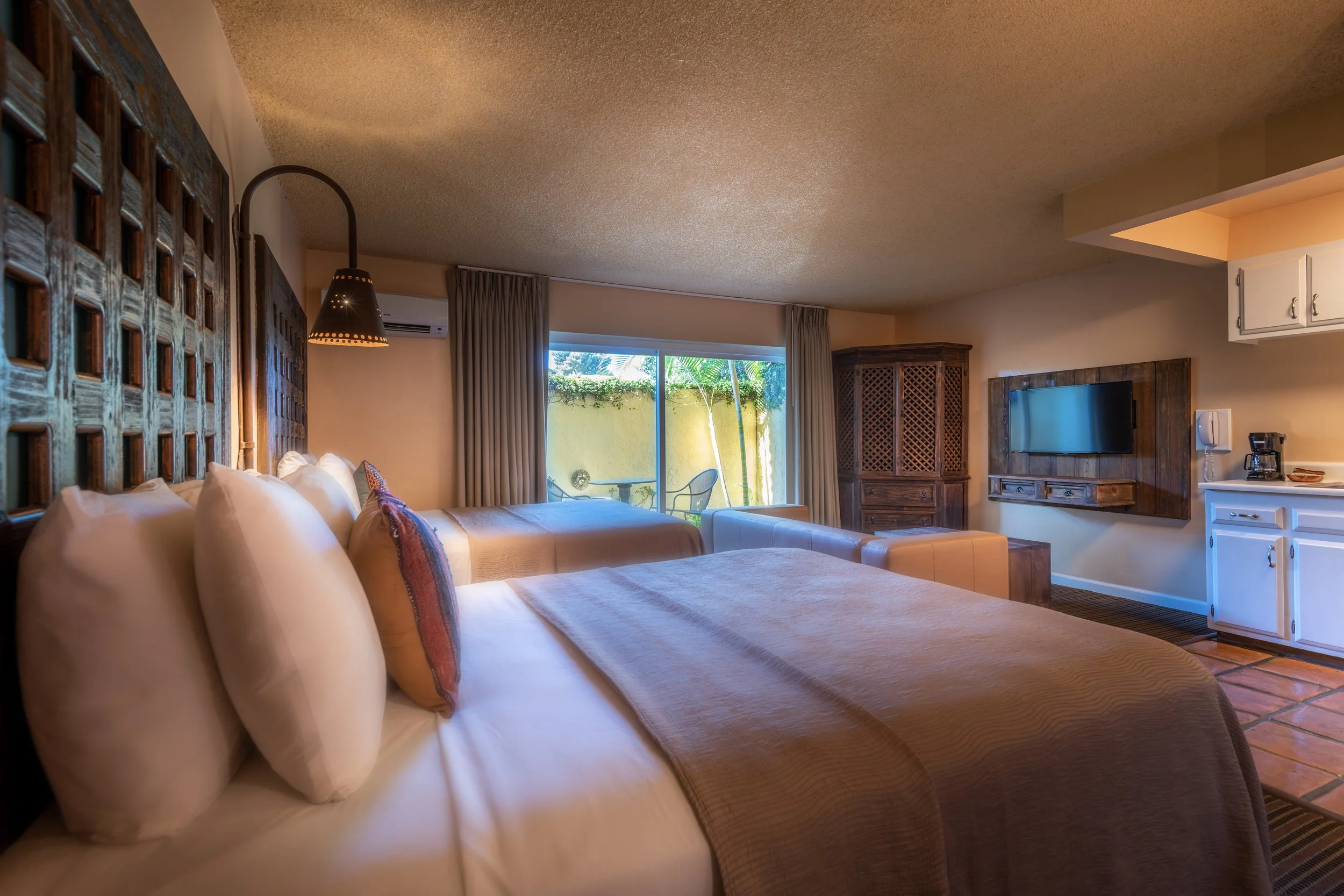 Hotel room with two neatly made beds, a large sliding glass door leading to a small outdoor patio, a wall-mounted flat-screen TV, a wooden cabinet, white kitchenette cabinets, and outdoor chairs.