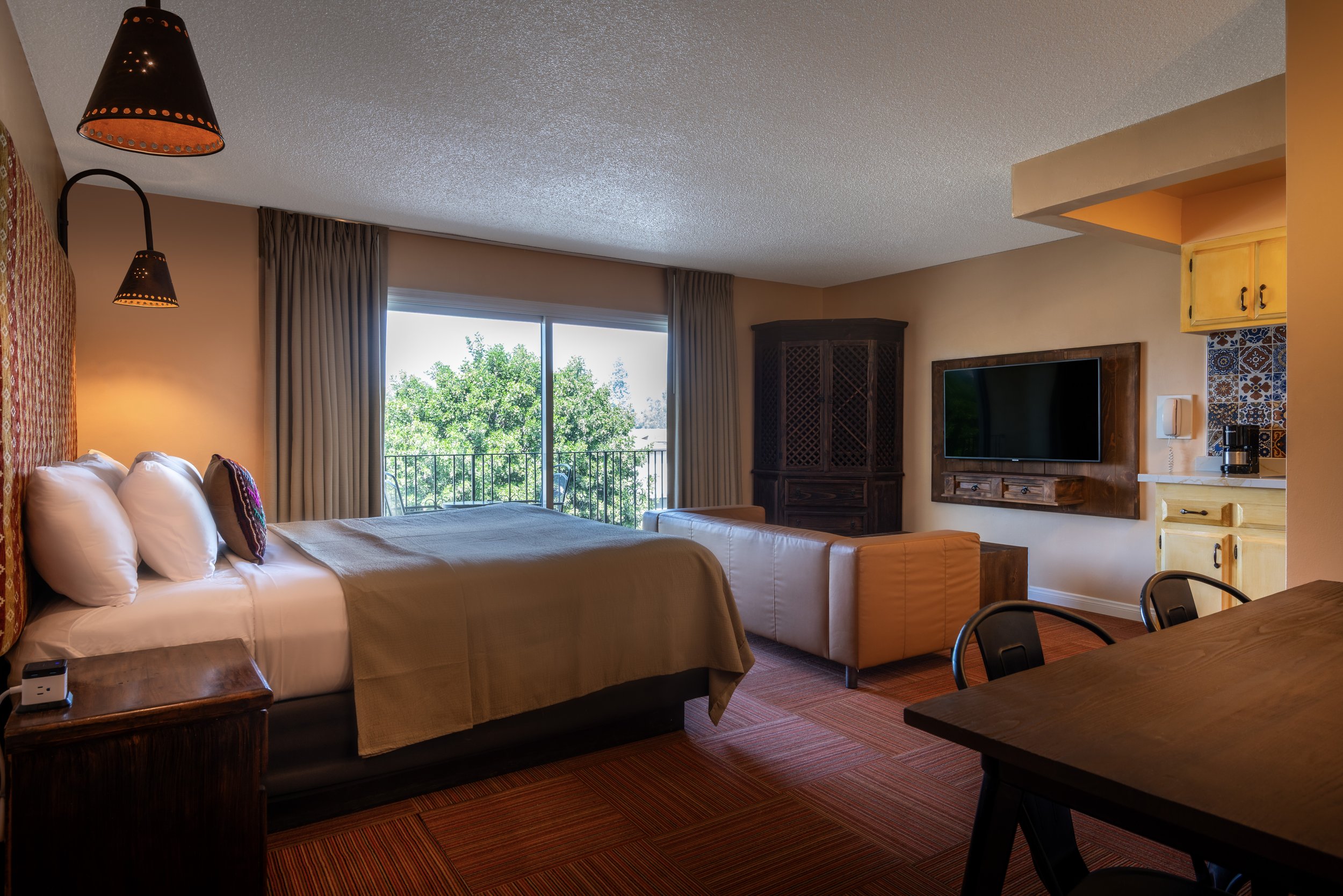 Hotel room with a bed, a couch, a large window, a television, and a kitchenette, decorated in warm colors.