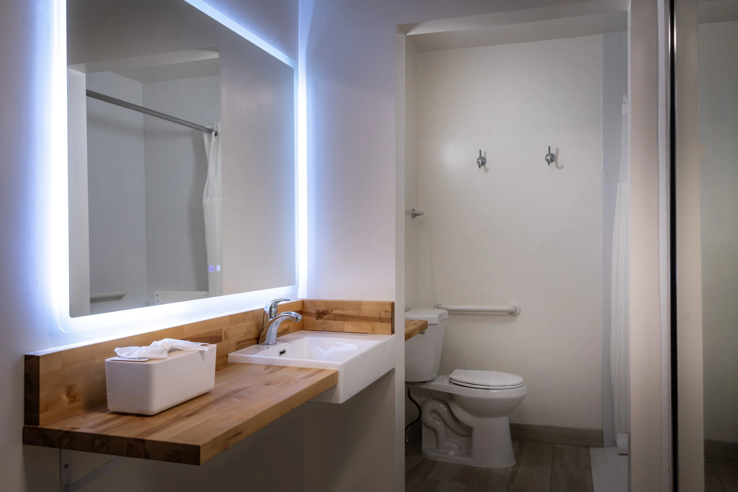 Bathroom with illuminated mirror, wooden countertop, sink, toilet, two hooks on wall, grab bar on wall, shower area with curtain
