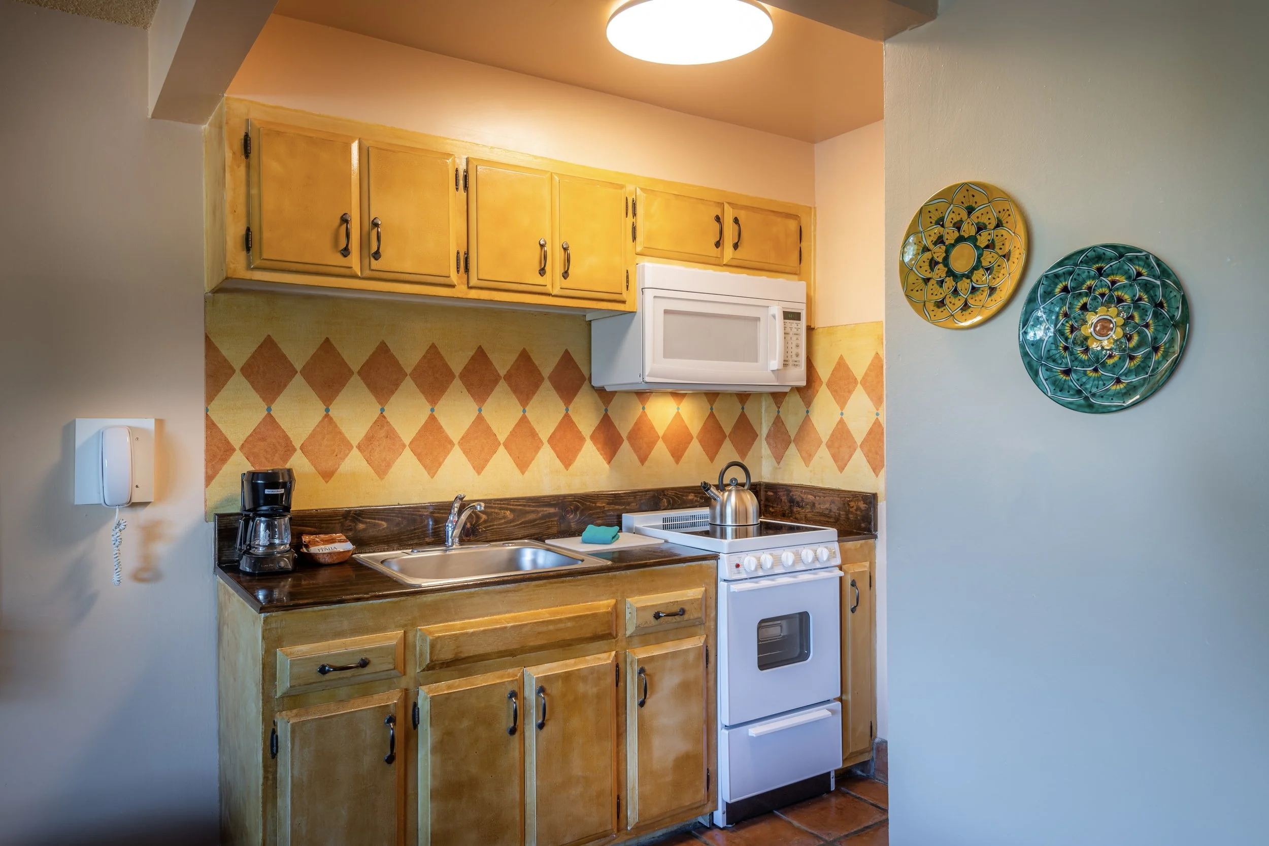 Small kitchen with yellow cabinets, a white microwave, a coffee maker, a sink, a stove with a teapot, and decorative plates on the wall.