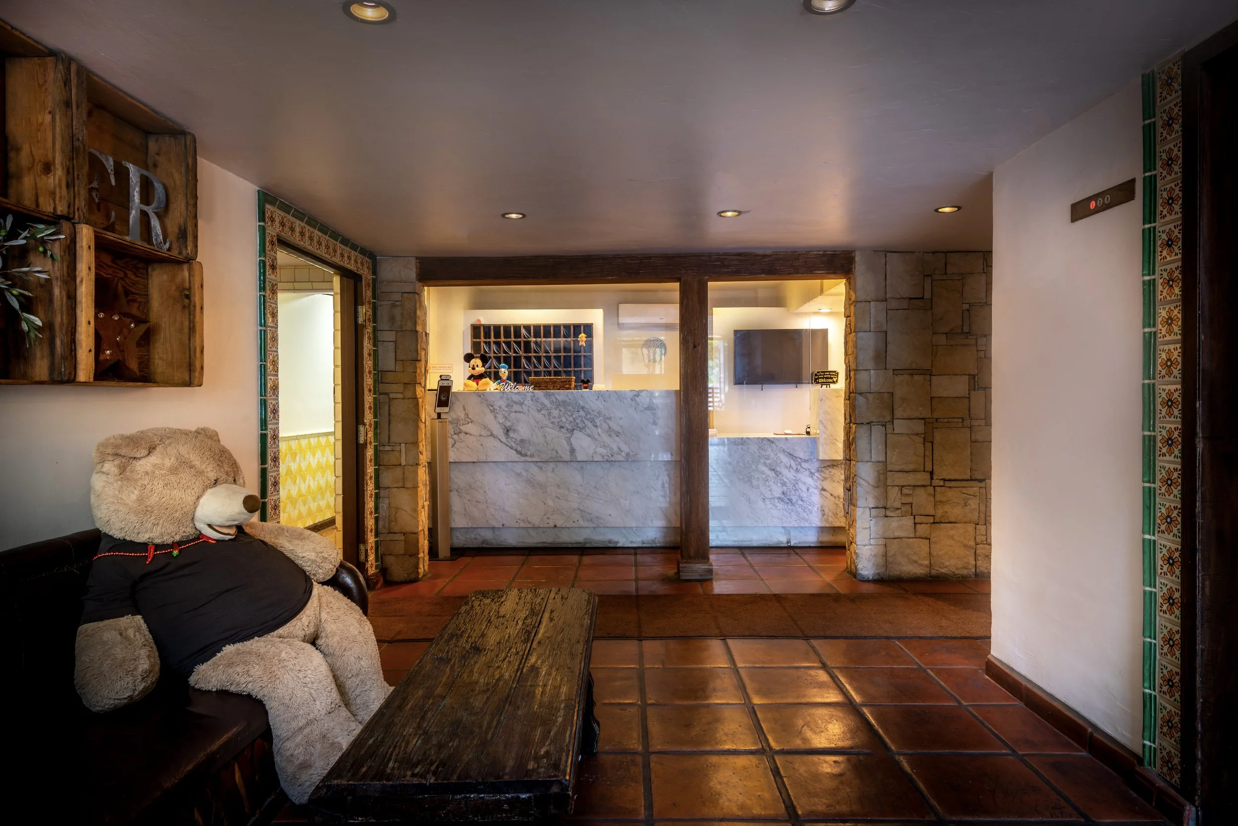 Hotel lobby with a large teddy bear sitting on a black leather couch, wooden coffee table in front, marble reception desk in the background ,and tiled floor.