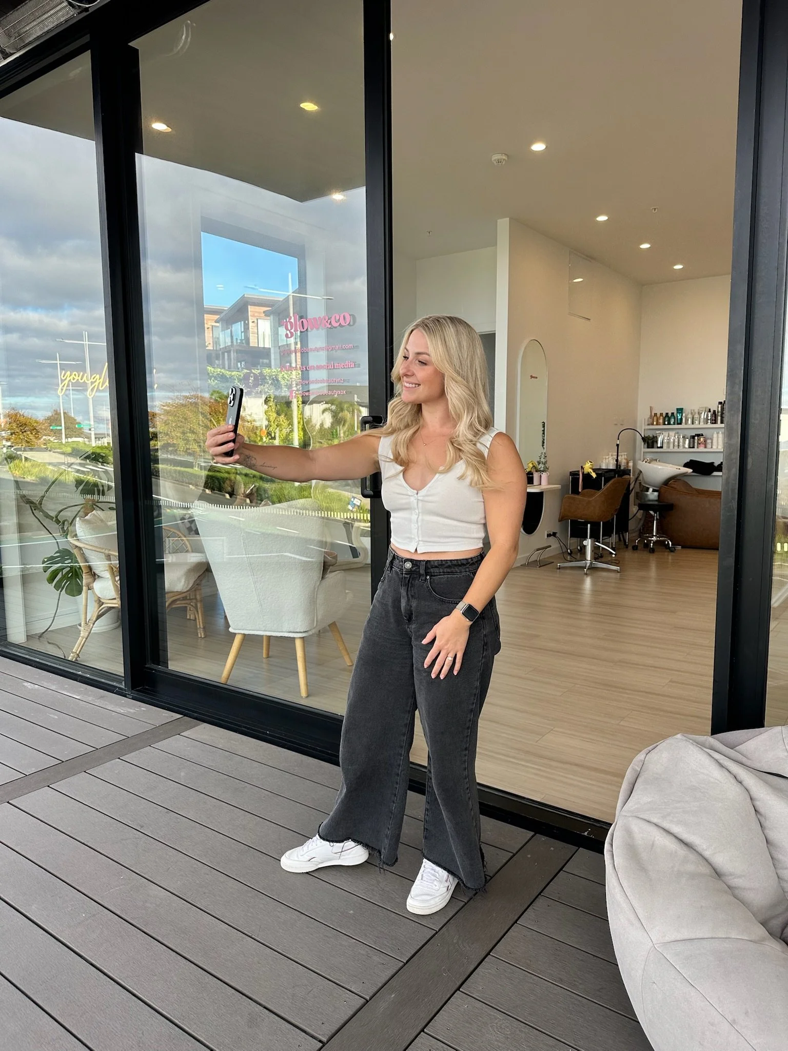 A woman standing on a wooden deck outside a modern glass-walled salon, taking a selfie with her phone. She has long blonde hair, a white sleeveless top, black wide-leg jeans, white sneakers, and is smiling. Inside the salon, mirrored stations and sty