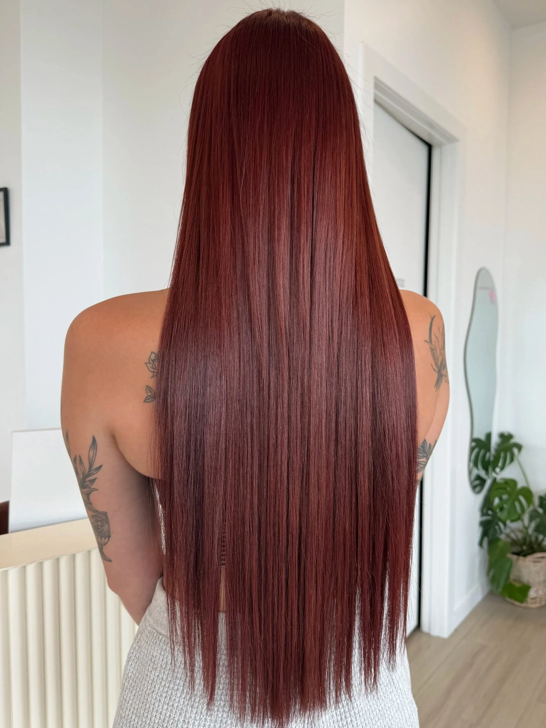 Back view of a woman with long, straight, vibrant red hair, showing tattoos on her arms, standing in a room with white walls, a mirror, and a potted plant.