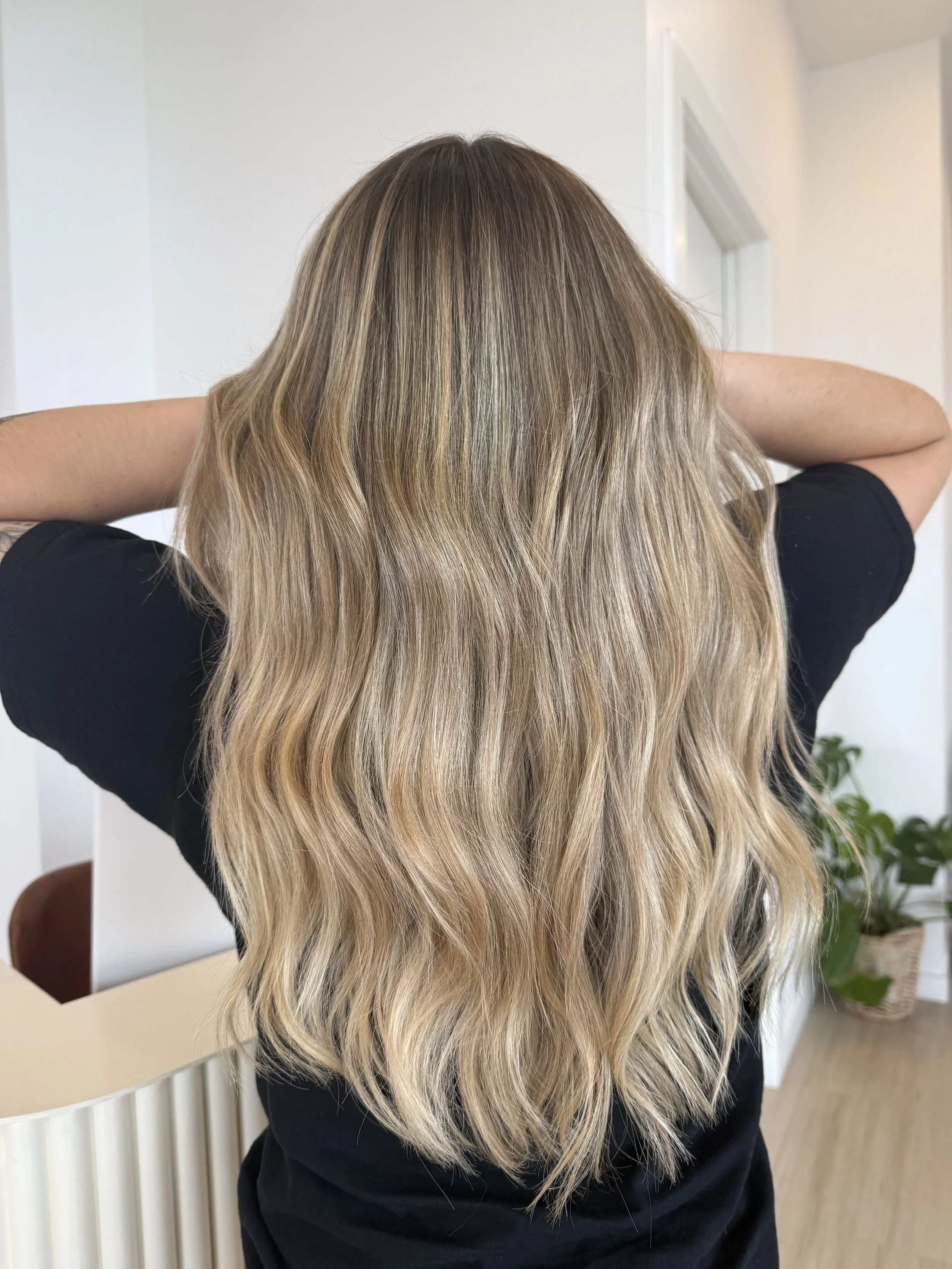 Woman with long, wavy blonde hair, wearing a black shirt, standing indoors with arms raised behind her head.
