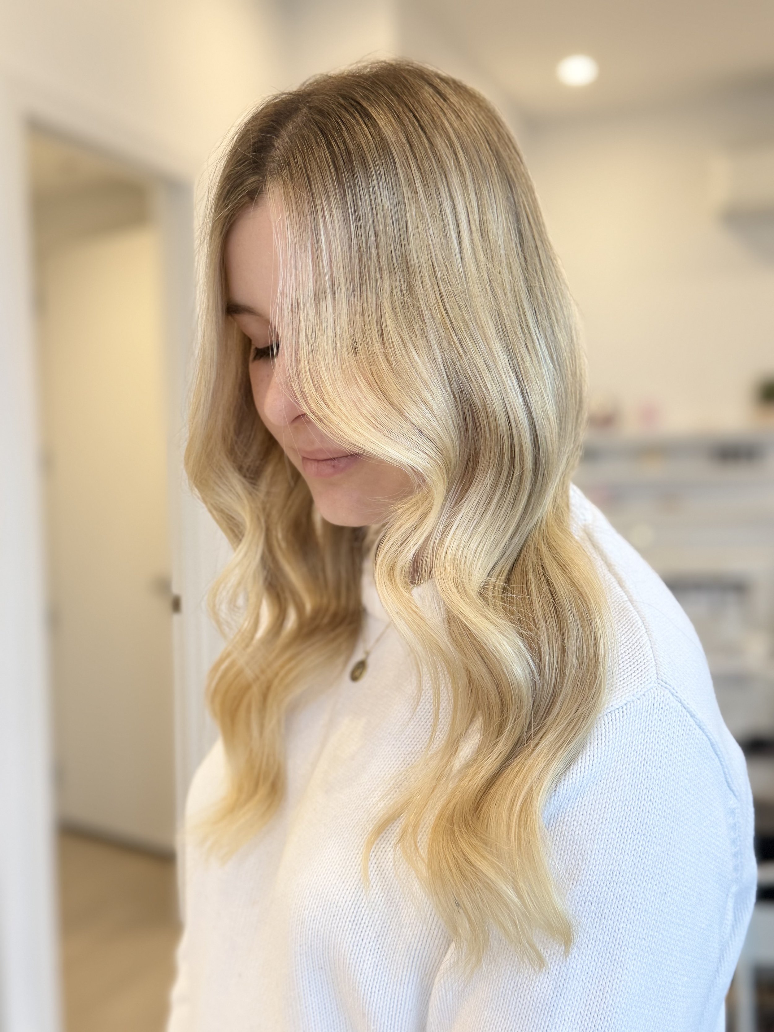 Woman with long, wavy blonde hair styled with loose waves, wearing a white top and a gold necklace, standing indoors with a blurred background.