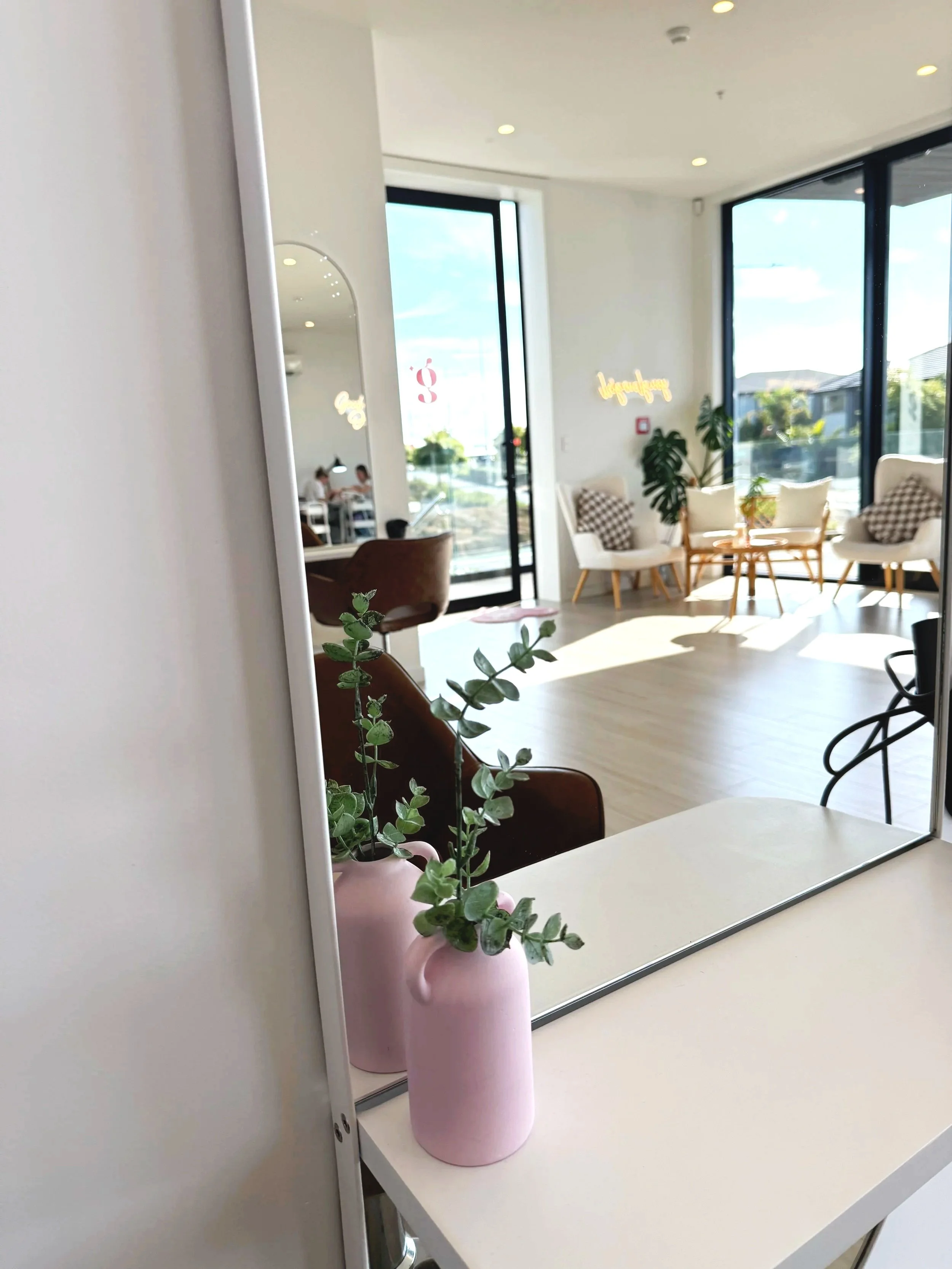 Interior of a hair salon with large windows, seating area with chairs and cushions, and a pink vase with green foliage on a white table.