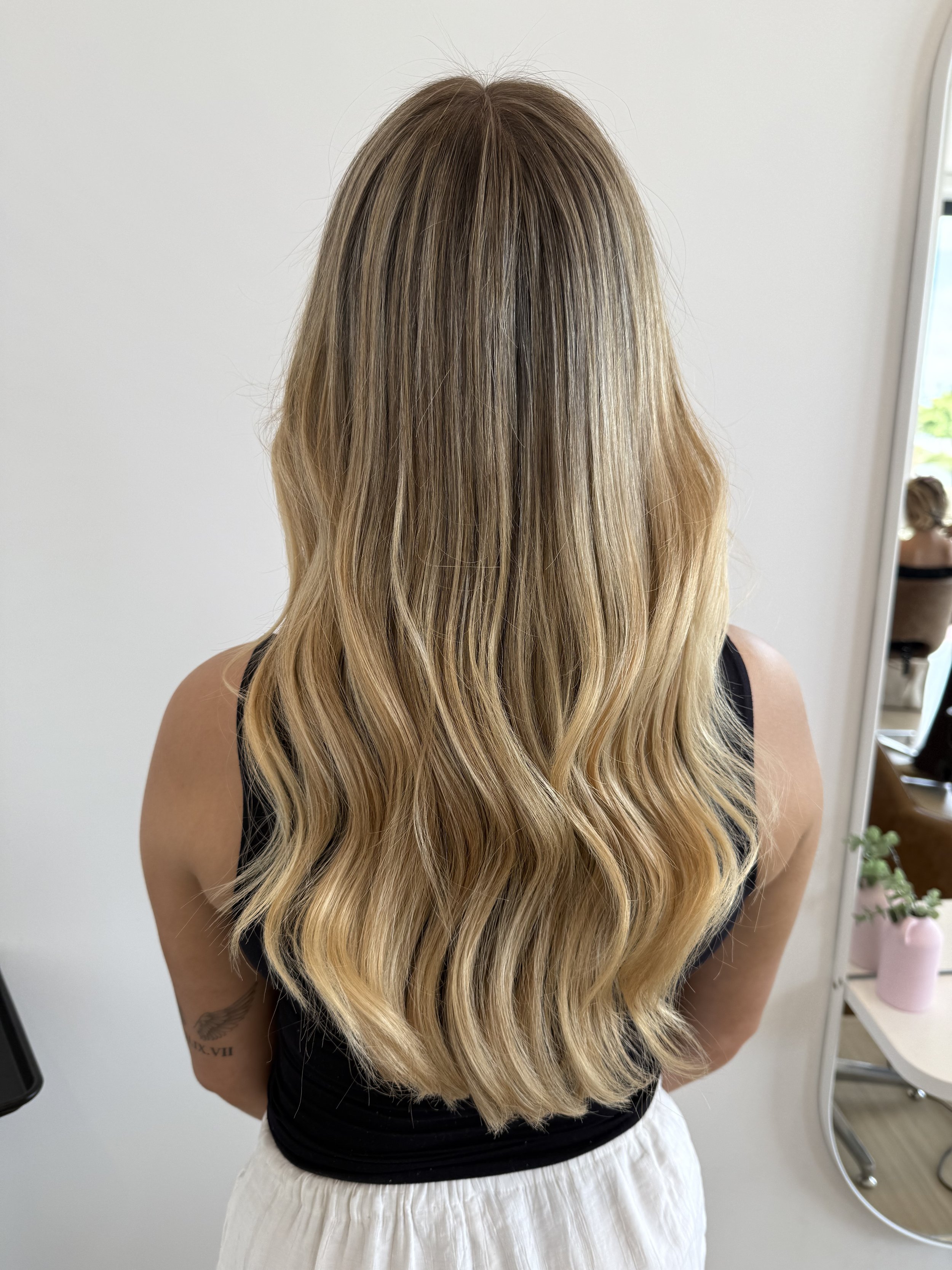 Back view of a woman with long, wavy, blonde hair standing indoors, near a mirror and white wall
