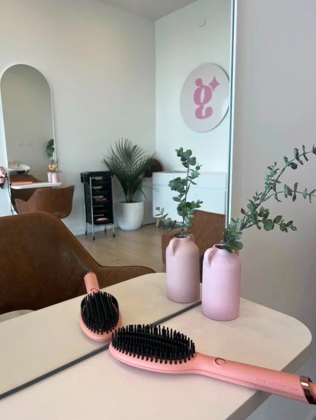 A salon interior with brown chairs, a mirror, and a white table holding two pink vases with green eucalyptus. On the table, there are two pink and black hairbrushes. In the background, a large green plant and a sign with a pink and white logo are vis