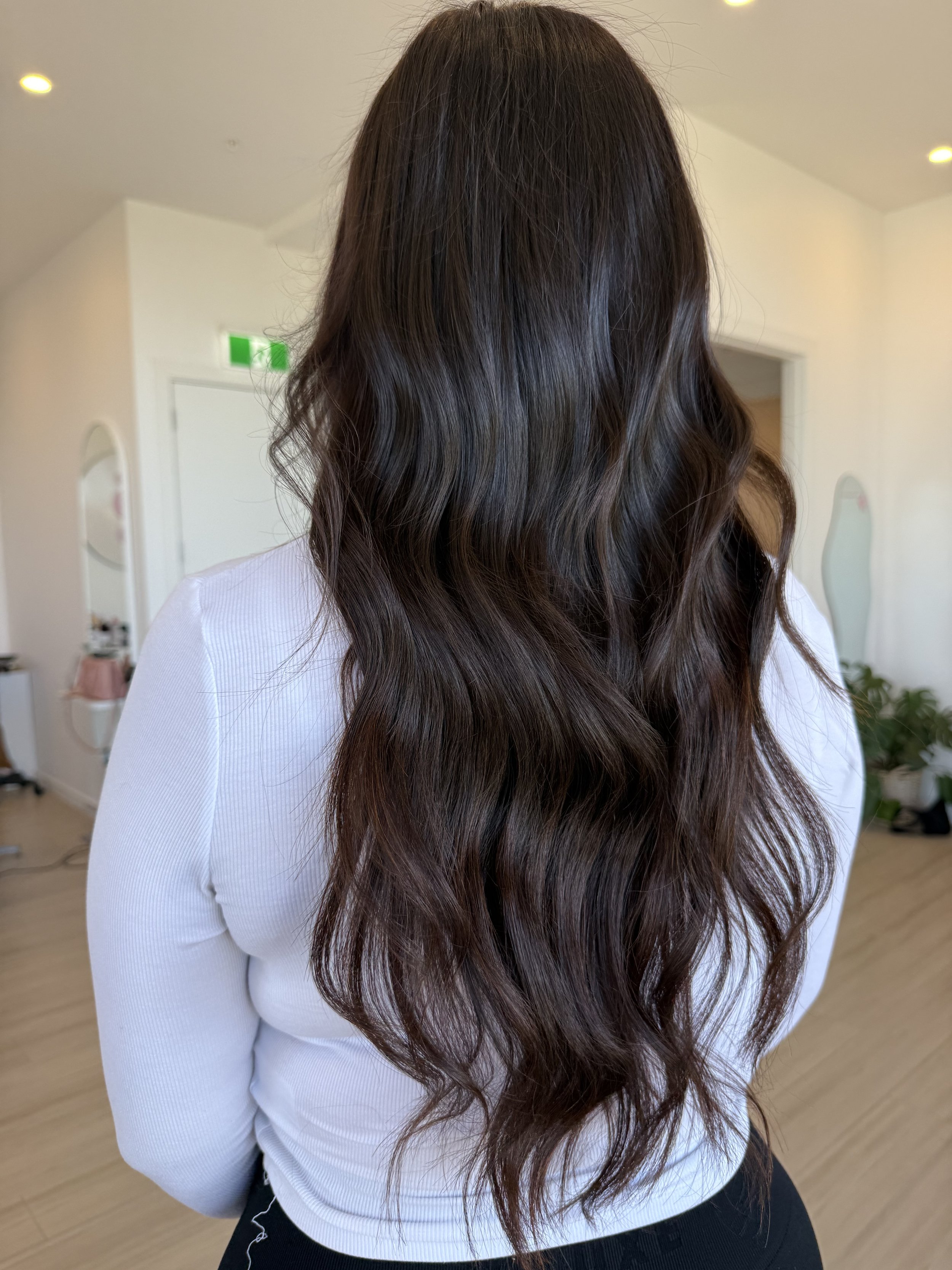 Back view of a woman with long, wavy brown hair wearing a white top in a brightly lit room.