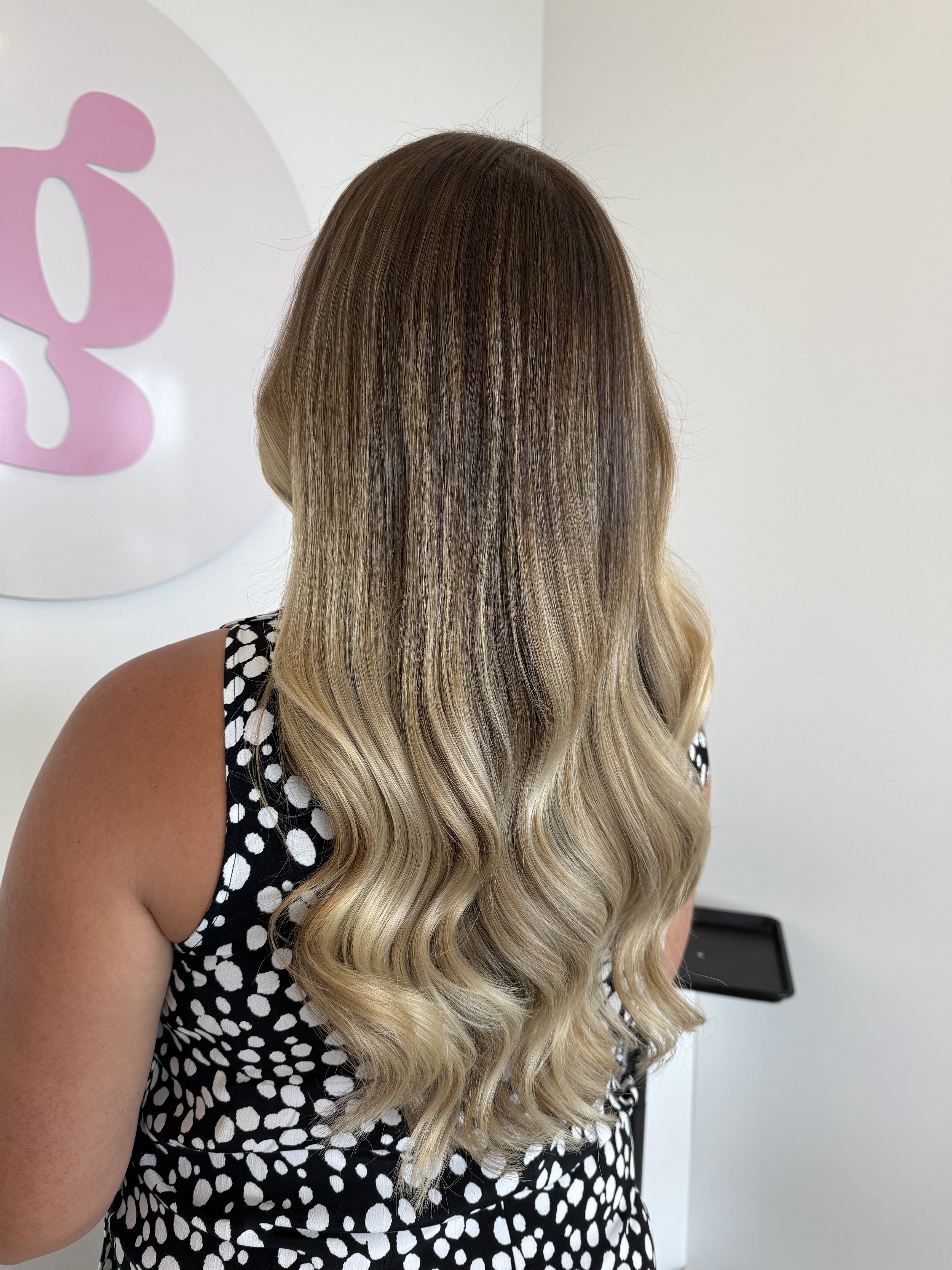 Back of a woman with long, wavy blonde hair, wearing a black dress with white polka dots, standing in front of a white wall with a pink and white logo.