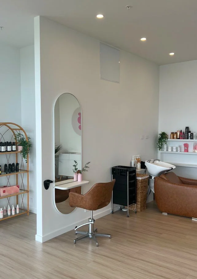 A modern salon interior with a white wall, round ceiling lights, a brown salon chair, a mirror, hair products on shelves, a hair wash basin, and a small black rolling cart.