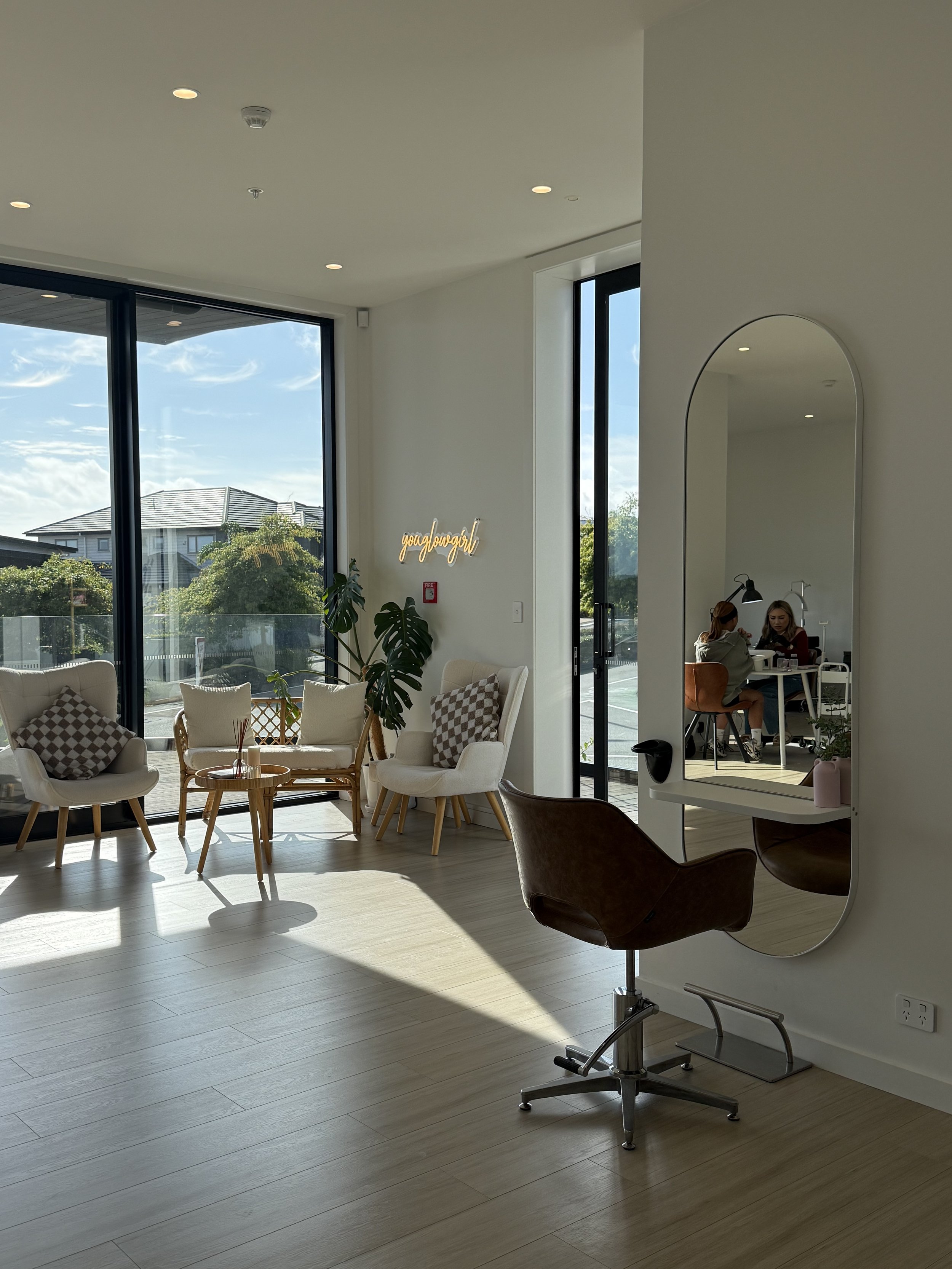 Interior of a modern, minimalistic salon or waiting area with white walls, large glass windows, a seating area with white armchairs, a small table, and a large mirror with a brown salon chair in front of it. Two women are sitting at a table in the ba