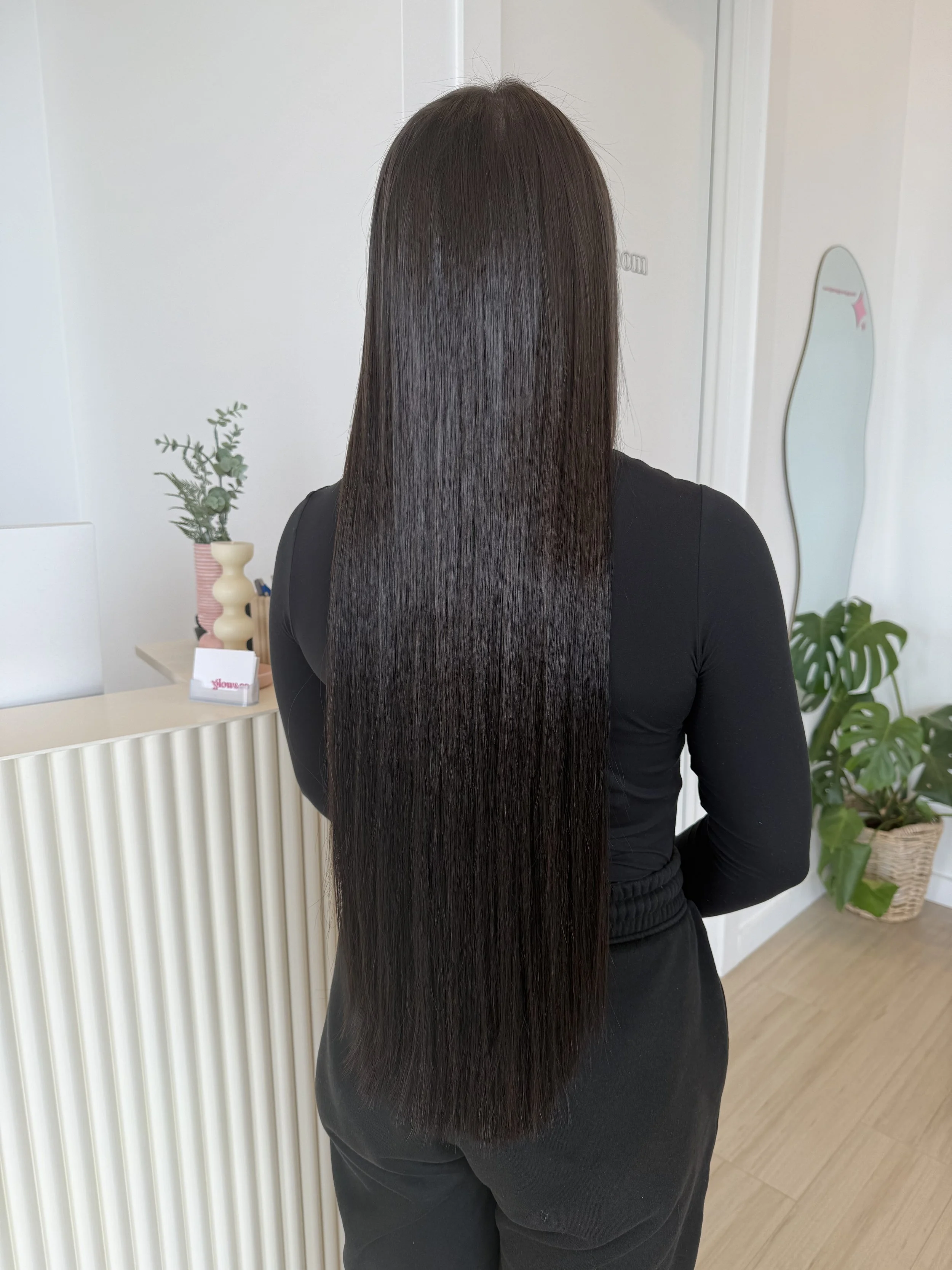Back view of a woman with long, straight, shiny brown hair wearing a black long-sleeve top in a modern interior space with plants and decorative items.