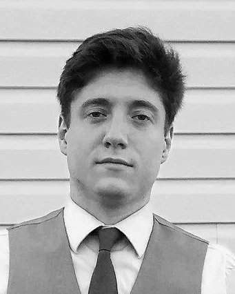 Black and white photo of a young man in a vest and tie standing in front of horizontal siding siding.