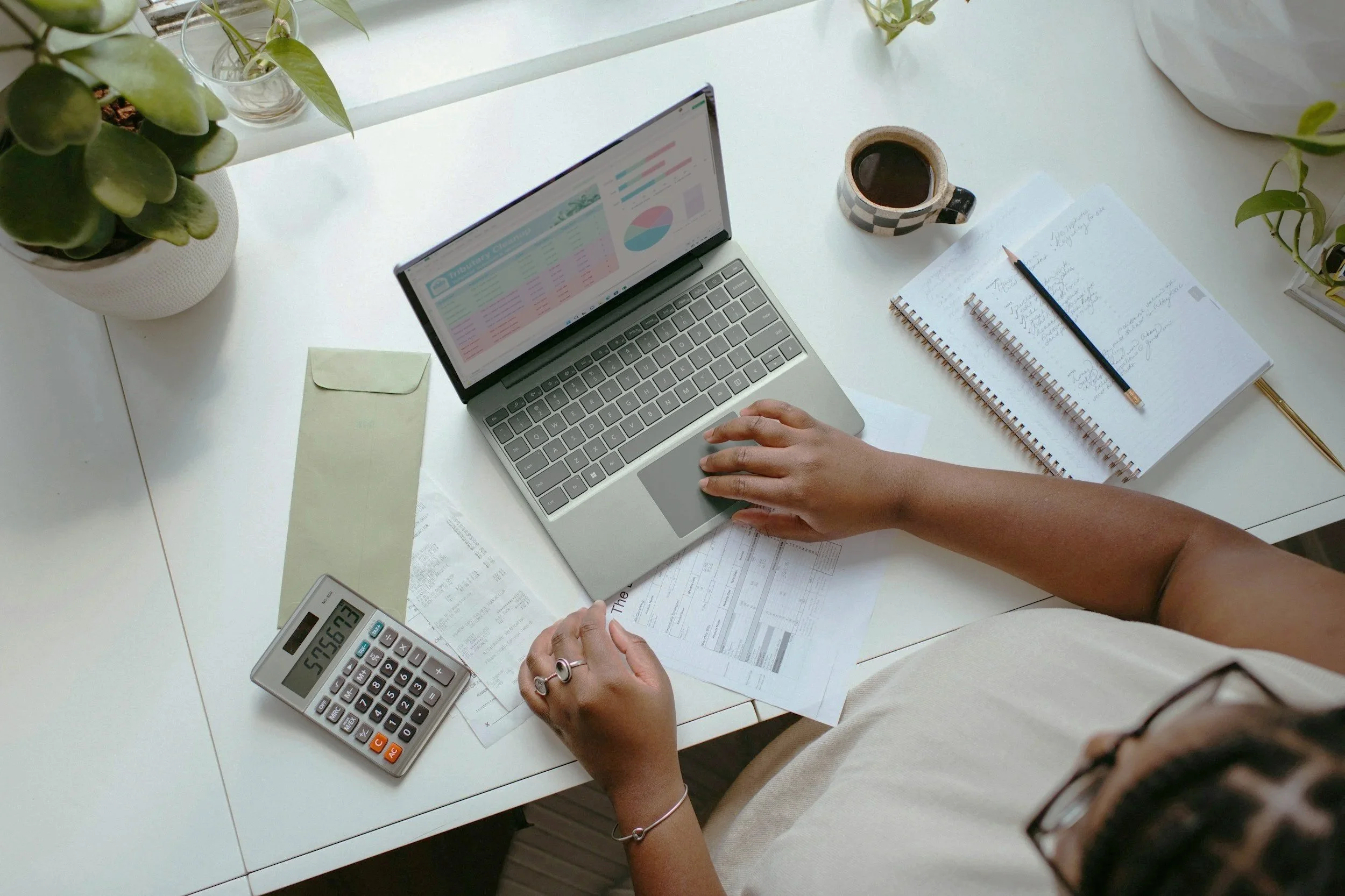 A person working at a white desk with a laptop, a calculator, notebooks, a pencil, a cup of coffee, and some documents. There are also two potted plants on either side of the desk.