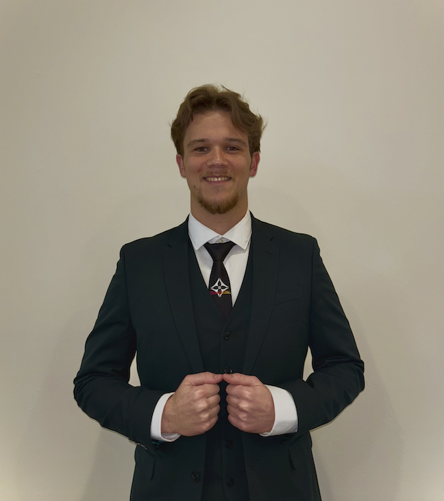 Young man in a black suit, white shirt, and black tie, smiling and standing against a plain light-colored wall.