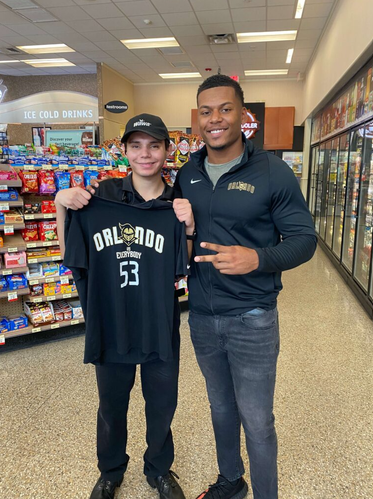 Two young men in a grocery store, one wearing a black Orlando basketball hoodie and the other wearing a black Orlando jacket, posing together and smiling. The man on the right is making a peace sign.