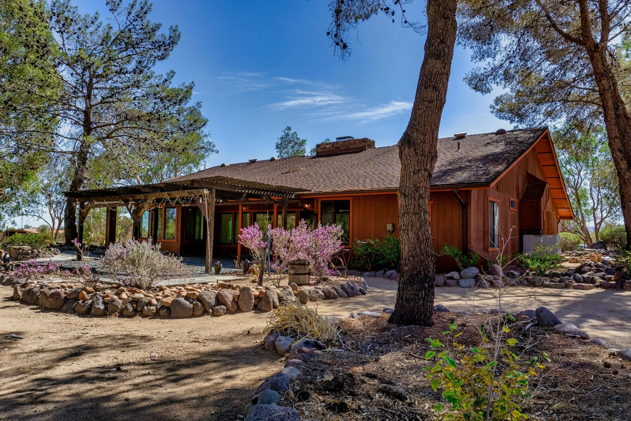 A single-story wooden house with a sloped roof, surrounded by trees and desert landscaping, including rocks, bushes, and flowering plants under a bright blue sky.