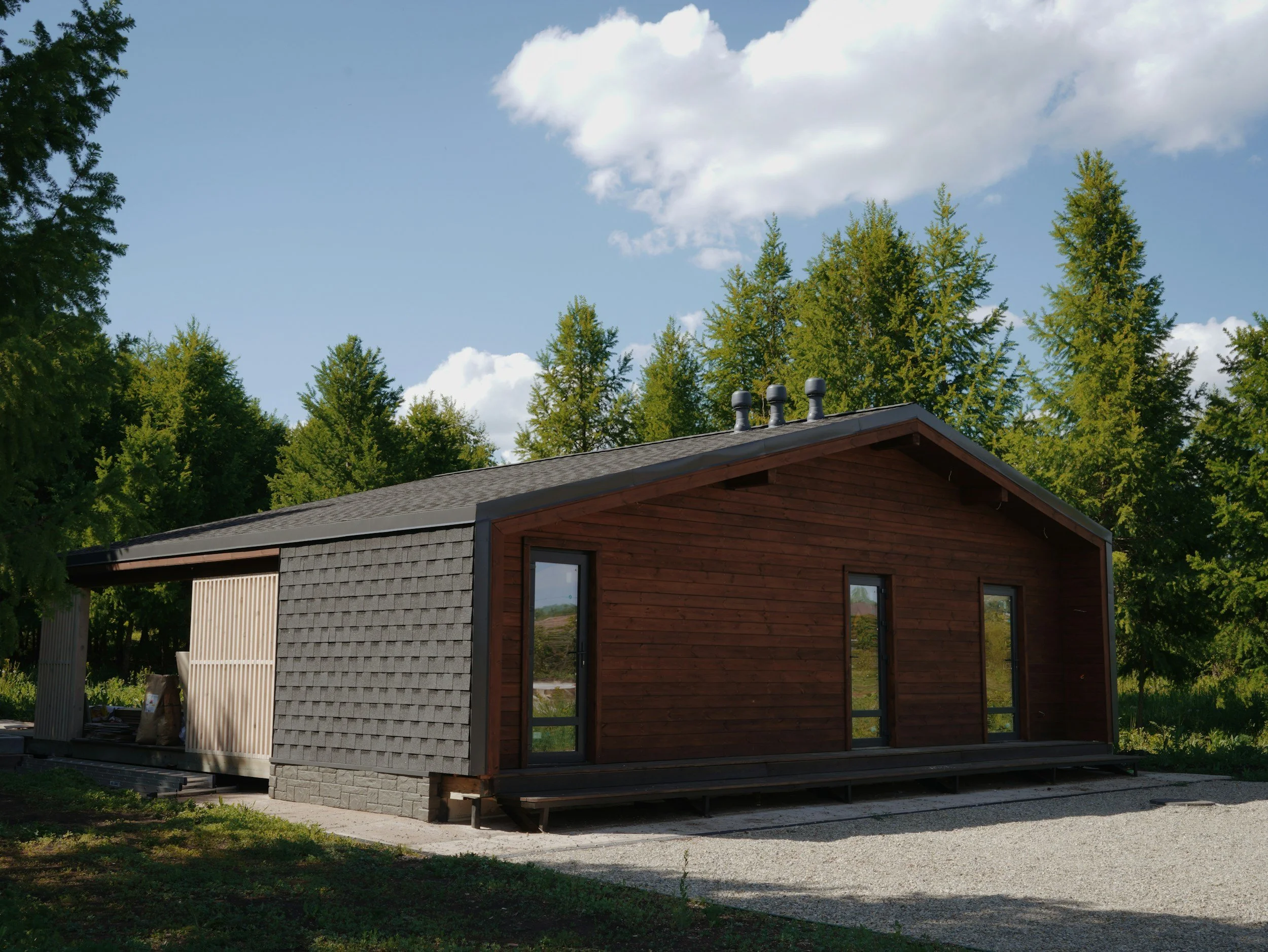 A modern house with a wooden and gray exterior, large glass doors, and a sloped roof, situated amidst greenery with tall trees and a partly cloudy sky.