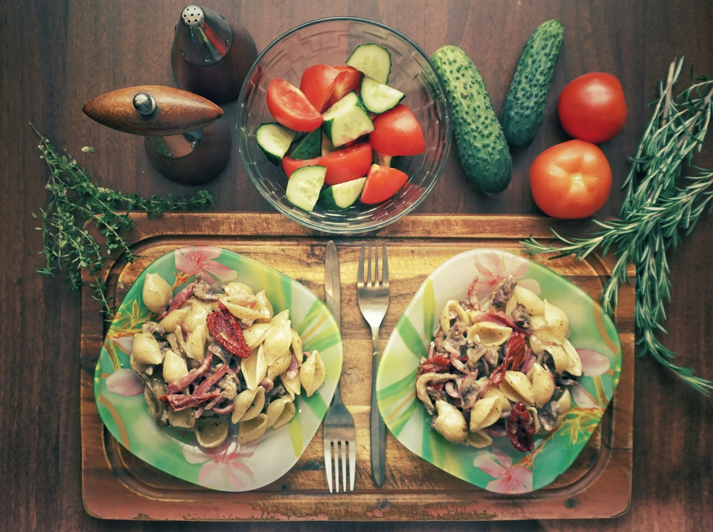 Overhead view of a meal with two plates of pasta with meat and sun-dried tomatoes, a bowl of chopped tomatoes and cucumbers, and fresh herbs like rosemary and thyme on a wooden tray.