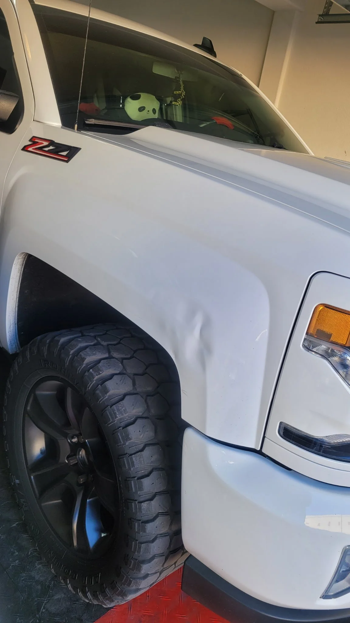Close-up view of the front part of a white Chevrolet Silverado Z71 truck, showing a dent on the fender, with a panda-shaped plush toy in the dashboard area seen through the windshield.