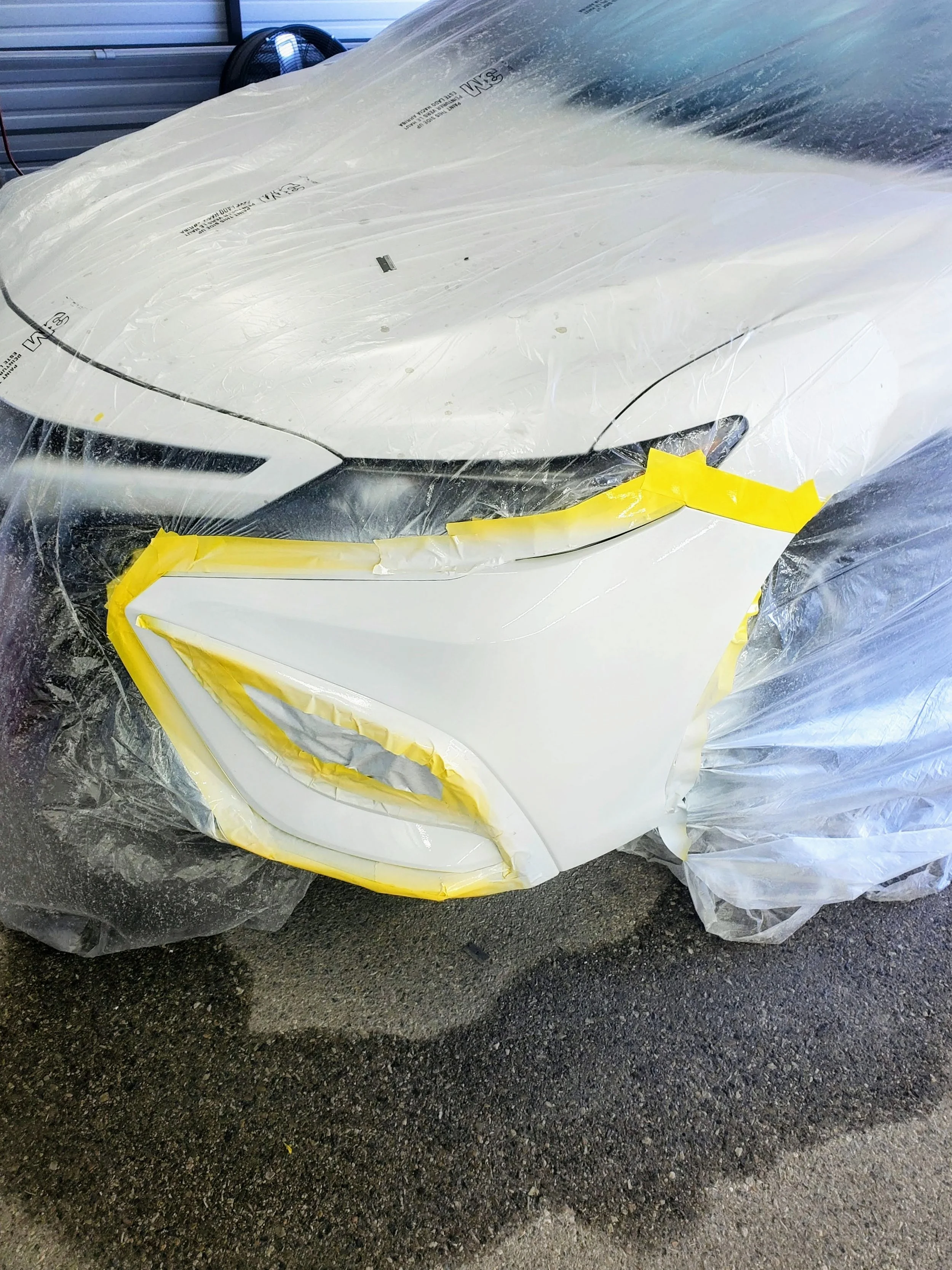 Close-up of a white car's front left corner with taped plastic covering for a paint job or repair, on a dark asphalt surface.
