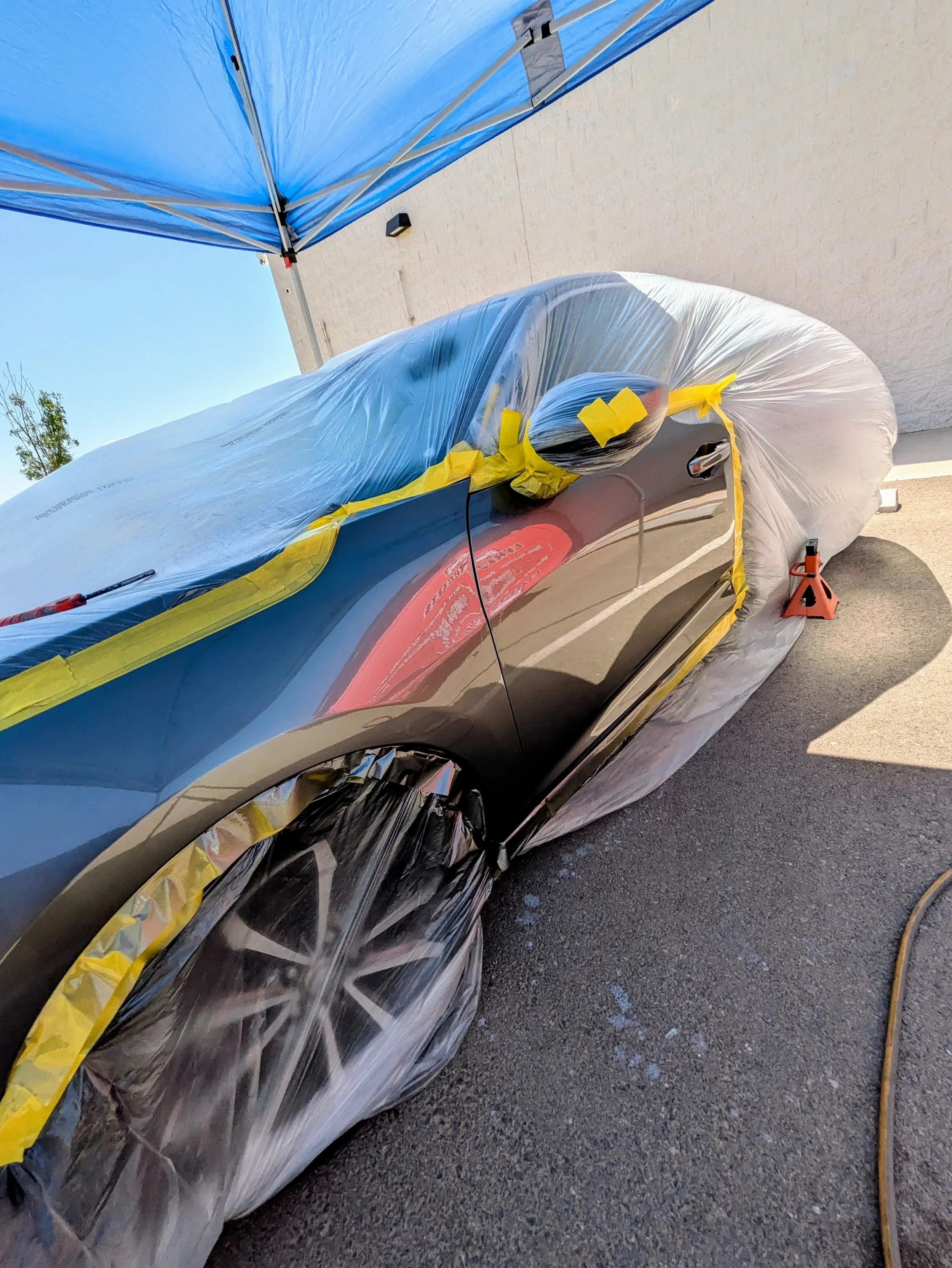 A black car covered with a clear plastic sheet and yellow tape, being prepared or protected for painting or maintenance outside under a blue canopy.