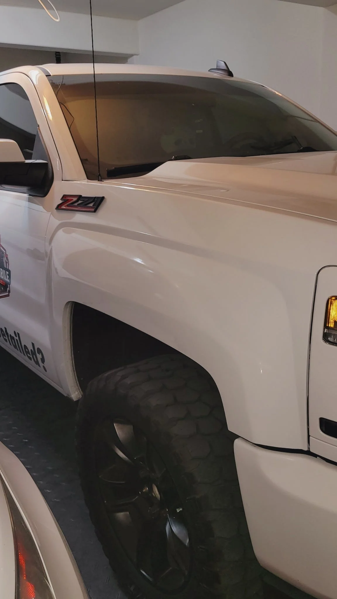 A white Chevrolet Silverado truck with Z71 off-road package parked indoors. The truck features a black and red Z71 badge on the front fender and a vehicle decal on the door. The truck has large, rugged black wheels with off-road tires.