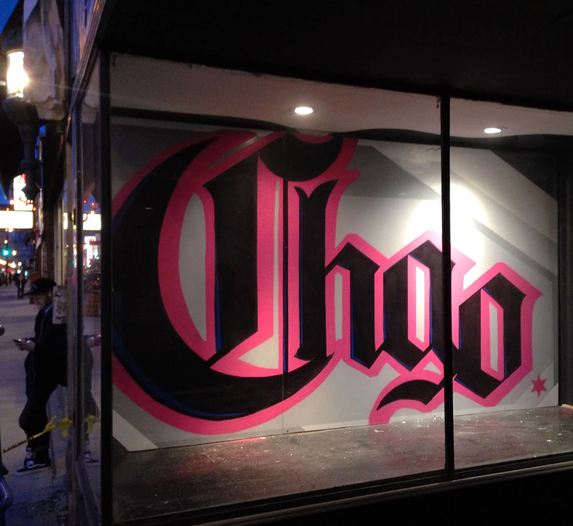 Street scene at night with graffiti art on a storefront window that spells 'Chgo' in black, pink, and white colors.