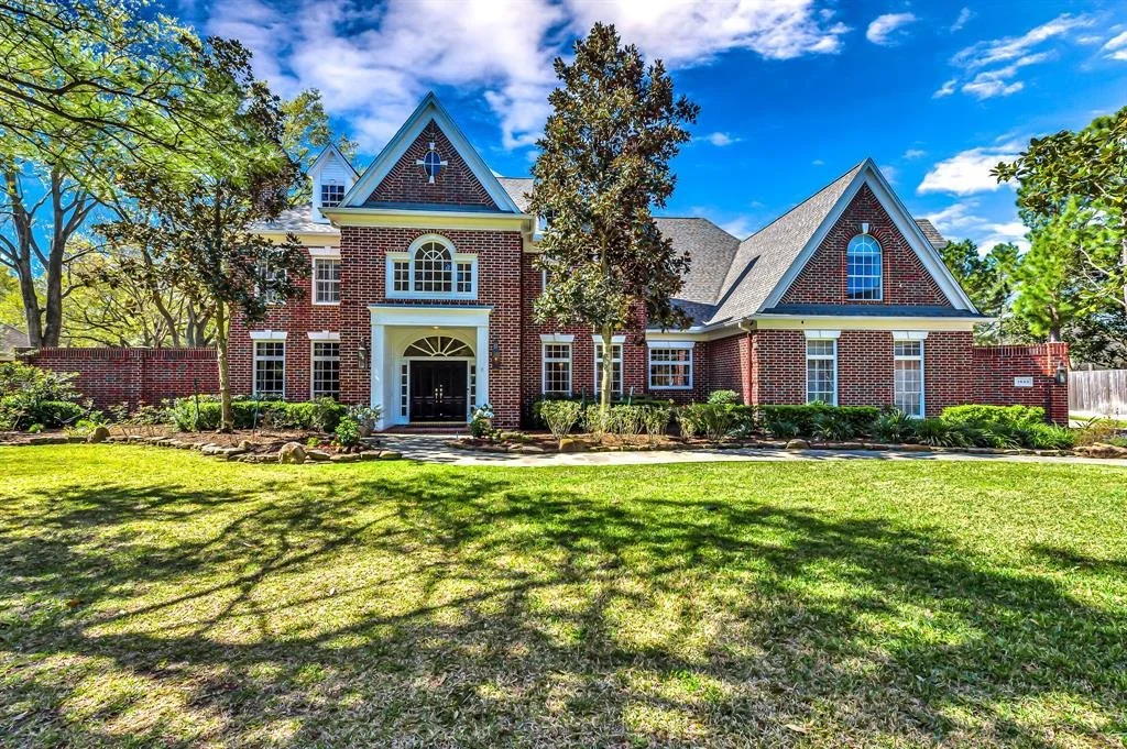 Large brick house with multiple peaked roofs, surrounded by trees and a well-maintained lawn under a partly cloudy sky.