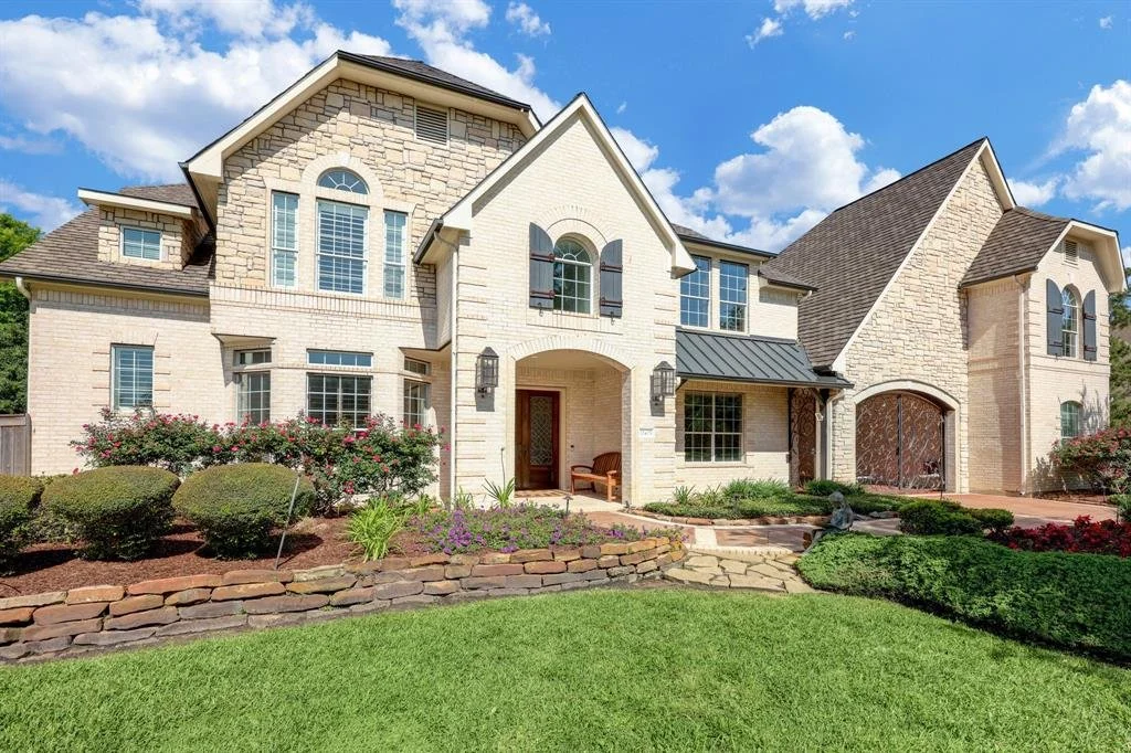 Large, elegant two-story house with a stone and brick exterior, multiple gabled roofs, tall windows, and a lush landscaped front yard with trimmed bushes, flowers, and a stone pathway.
