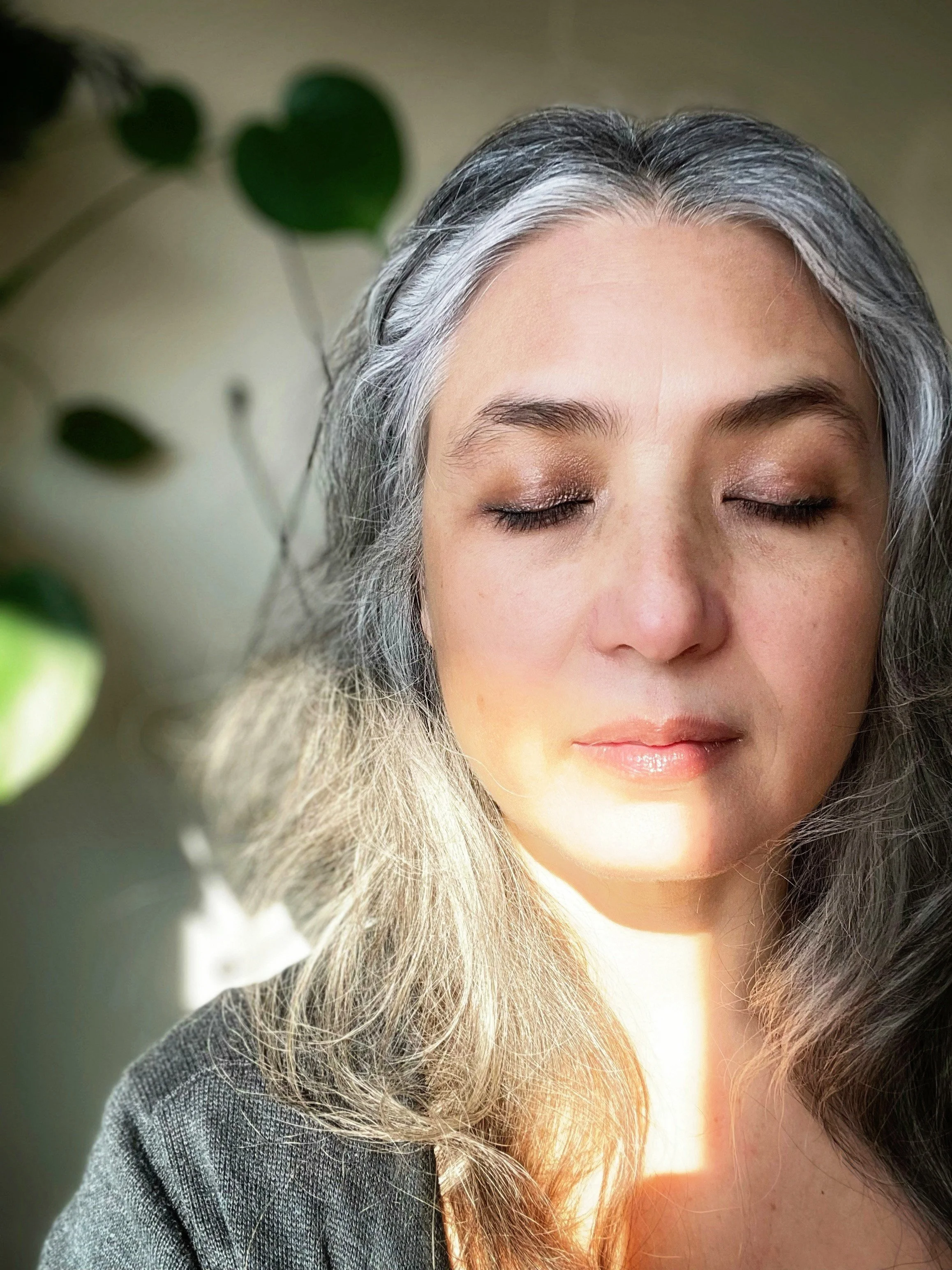 Close-up of a woman with closed eyes, gray and white hair, and a gentle expression, with sunlight illuminating her face and a blurred green plant in the background.