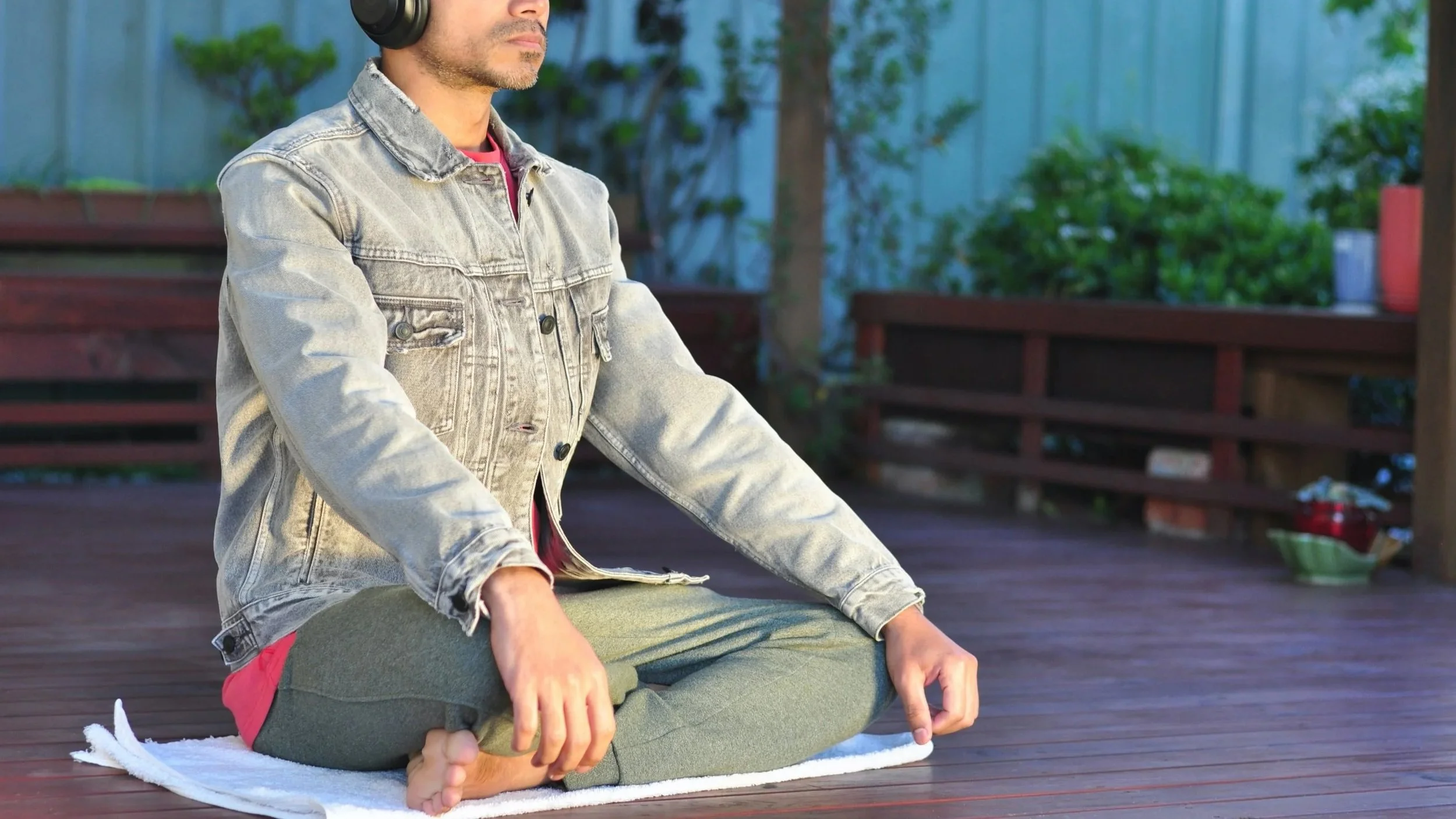 A person practicing yoga outdoors on a wooden deck, sitting cross-legged with hands resting on knees, wearing a denim jacket and headphones.