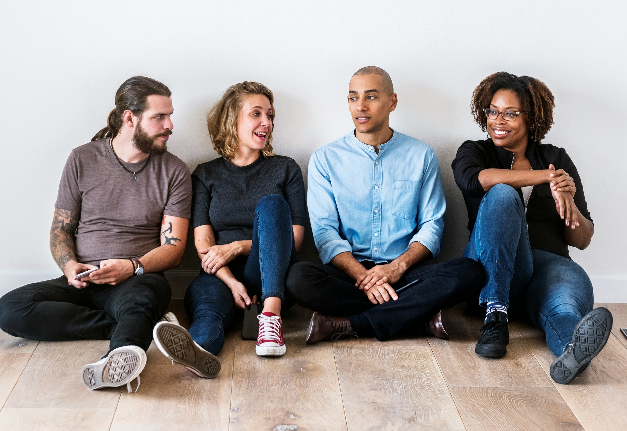 Group of four diverse young adults sitting on the floor against a white wall, engaged in conversation.