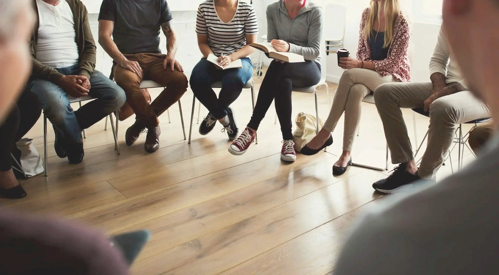 Group of people sitting in a circle during a discussion or meeting in a bright room with wooden floors.
