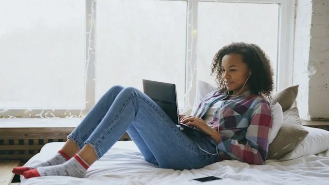 Young woman lying on bed using a laptop, wearing casual clothes and socks, in a bright room with large windows.