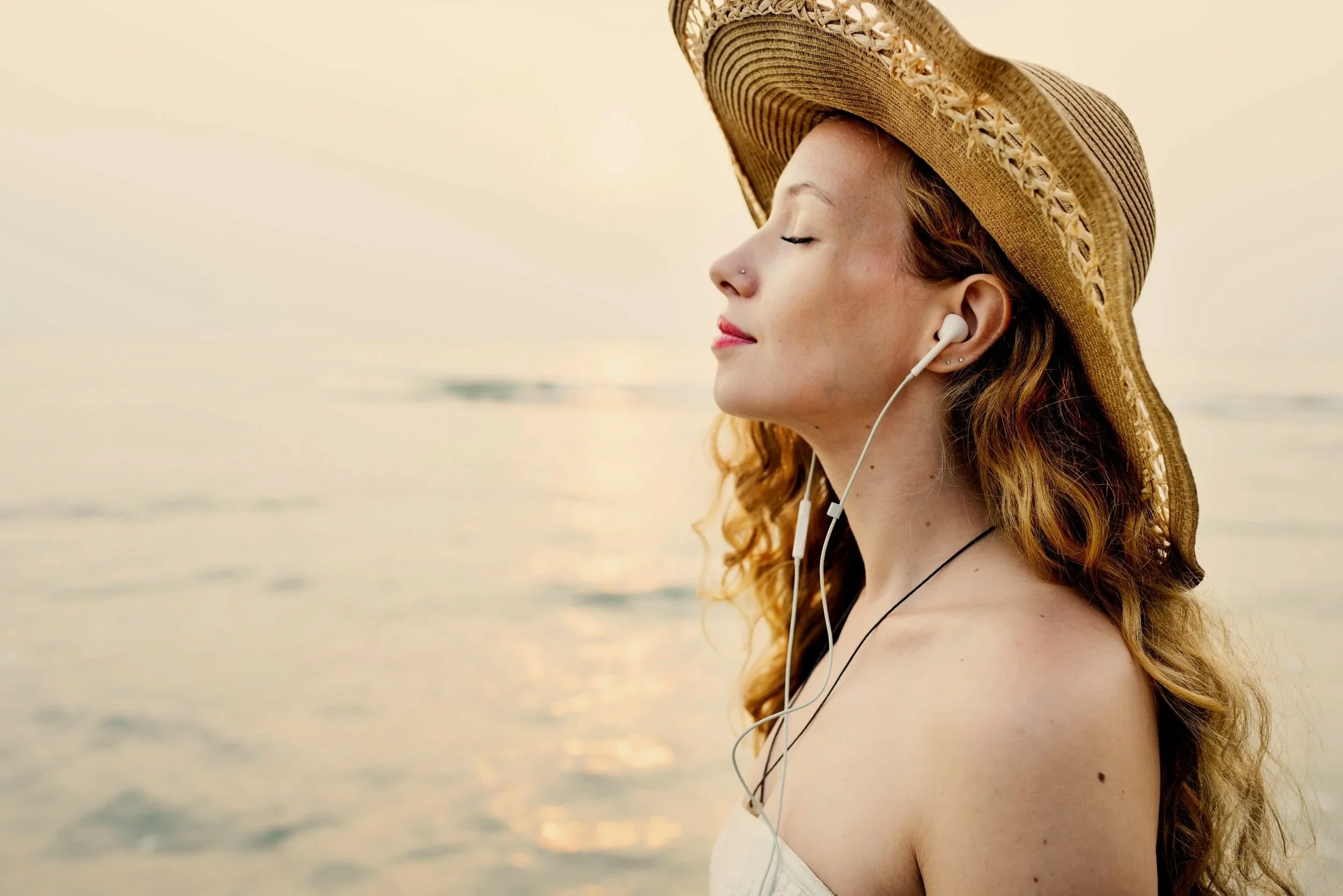 A young woman with red hair and closed eyes wearing a large straw sunhat and white earphones, standing outdoors near a body of water during sunset.