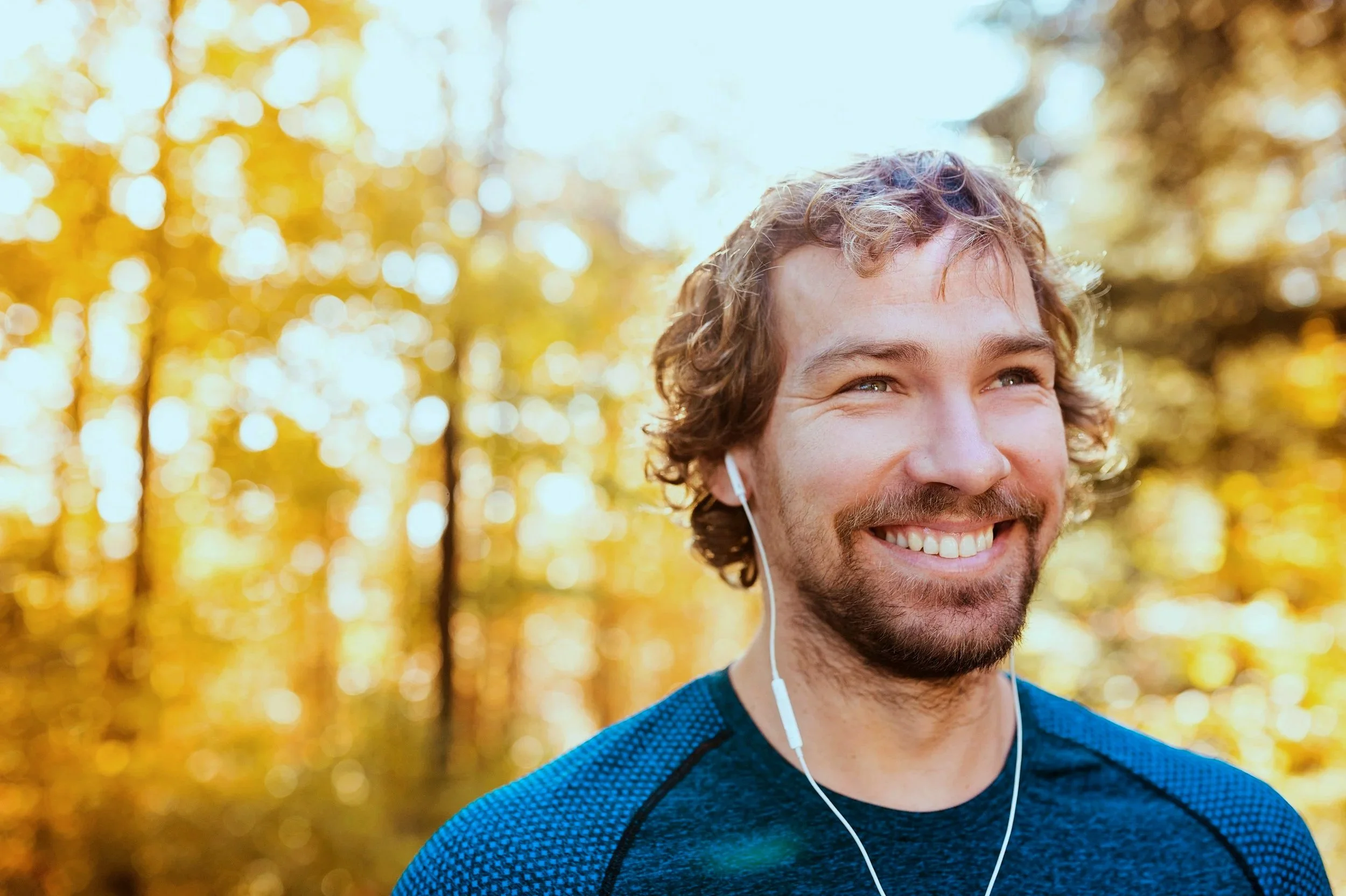 A smiling young man with curly hair, wearing a blue sports shirt, listening to music with white earphones outdoors in a park with golden autumn leaves.