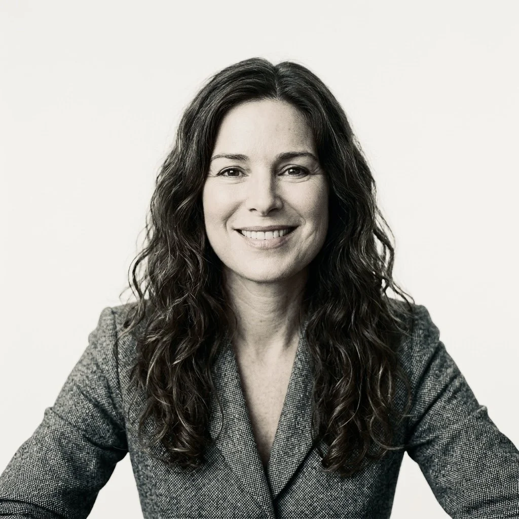 Portrait of a woman with wavy dark hair, wearing a gray blazer, smiling against a plain light background.