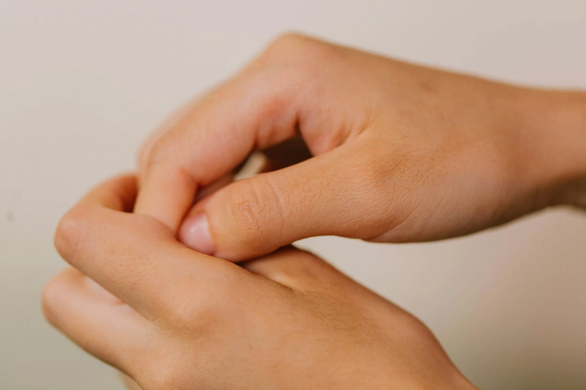 Close-up of a person's hand with fingers gently holding the thumb, with a plain, light background.