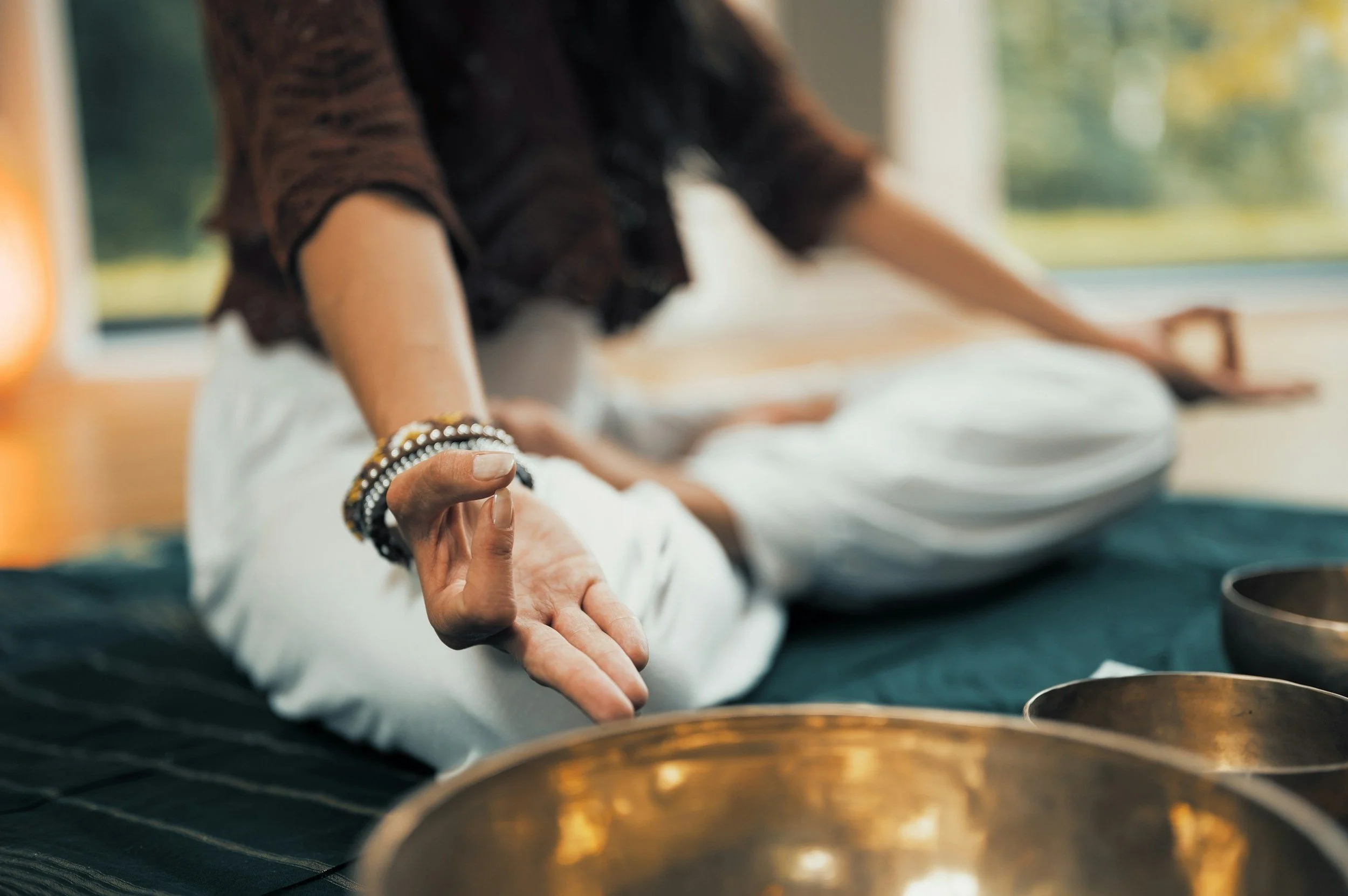 A person meditating with their eyes closed, sitting cross-legged on the floor, with their hand positioned in a mudra. The scene appears peaceful and indoors with natural light coming through a window.