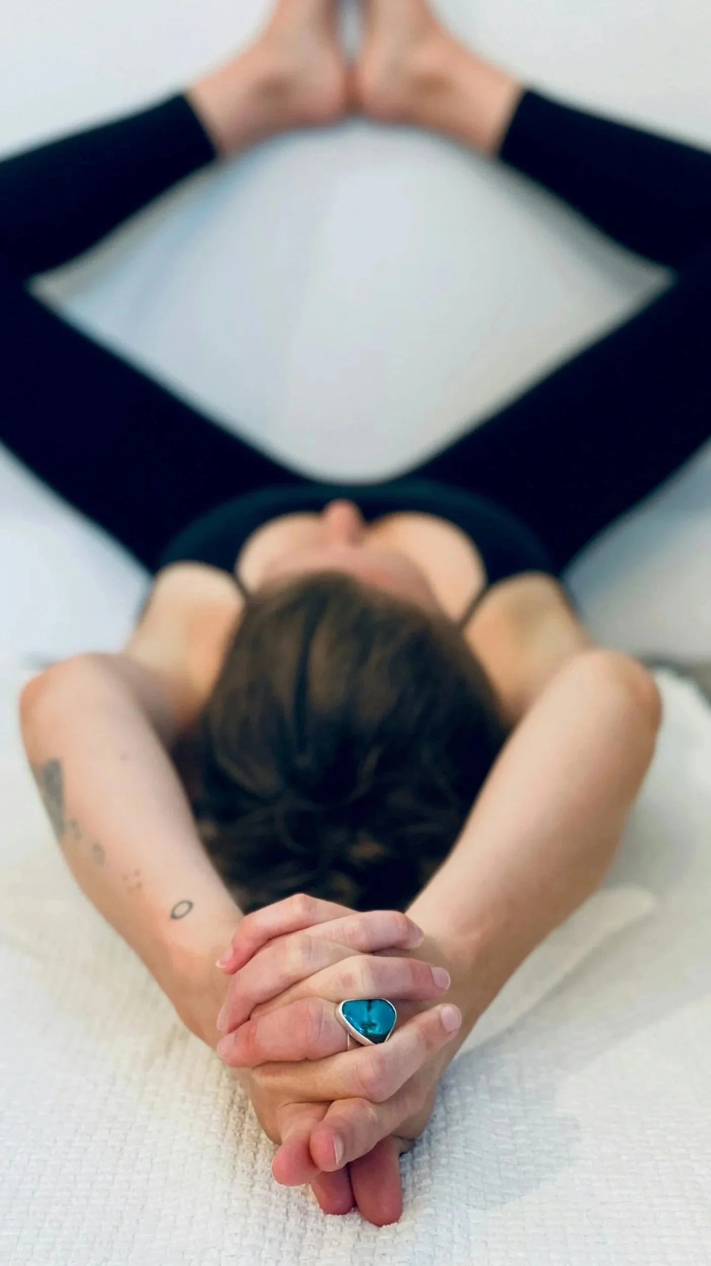 A woman lying on her back on a white surface with her eyes closed, arms resting behind her head, wearing a black sleeveless top, with tattooed arms and a large blue ring on her finger.